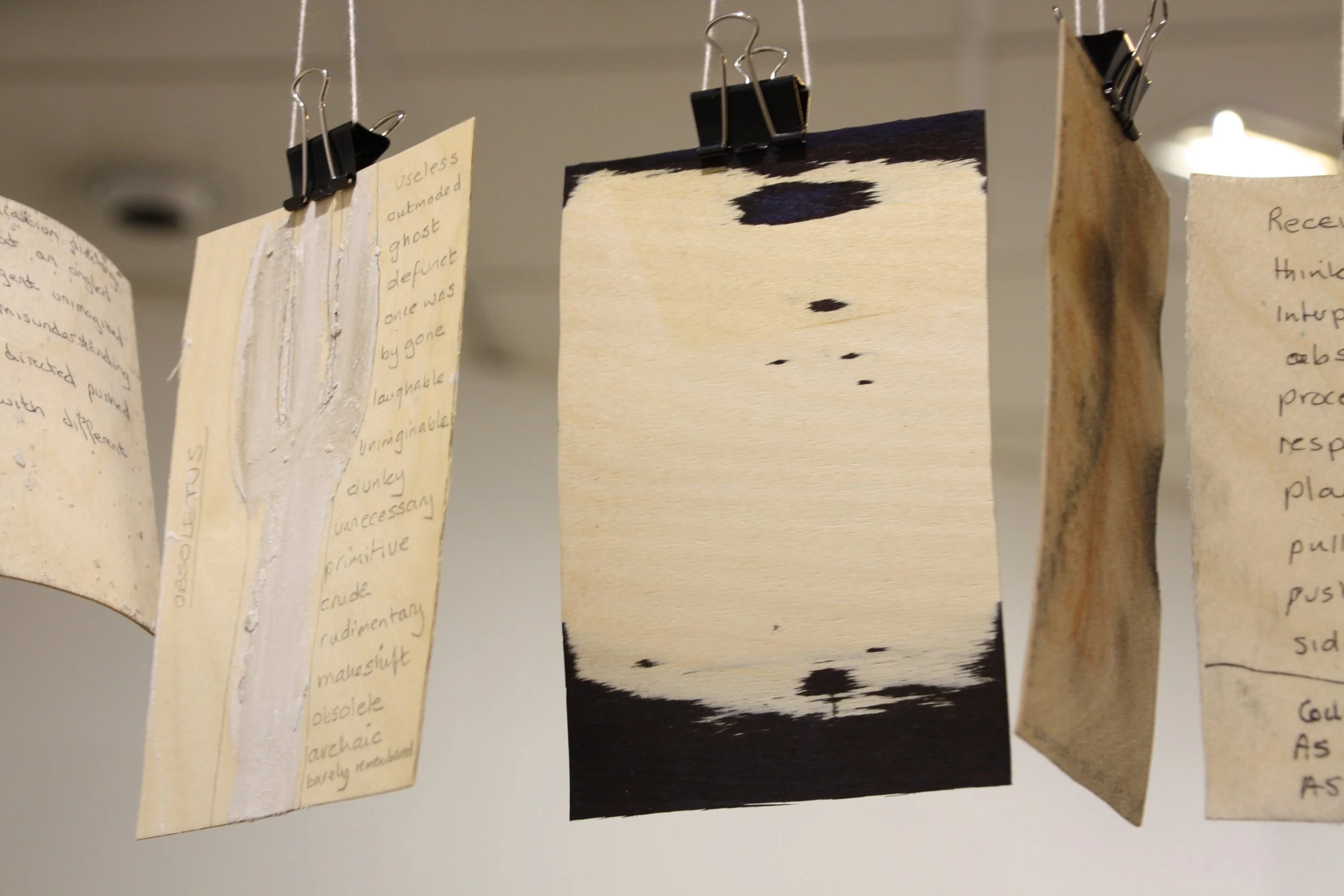 A series of handwritten notes on pieces of paper, clipped and hanging on strings with black binder clips. The visible notes contain scattered words and phrases, some of which are partially obscured, with a mix of cursive and print handwriting.