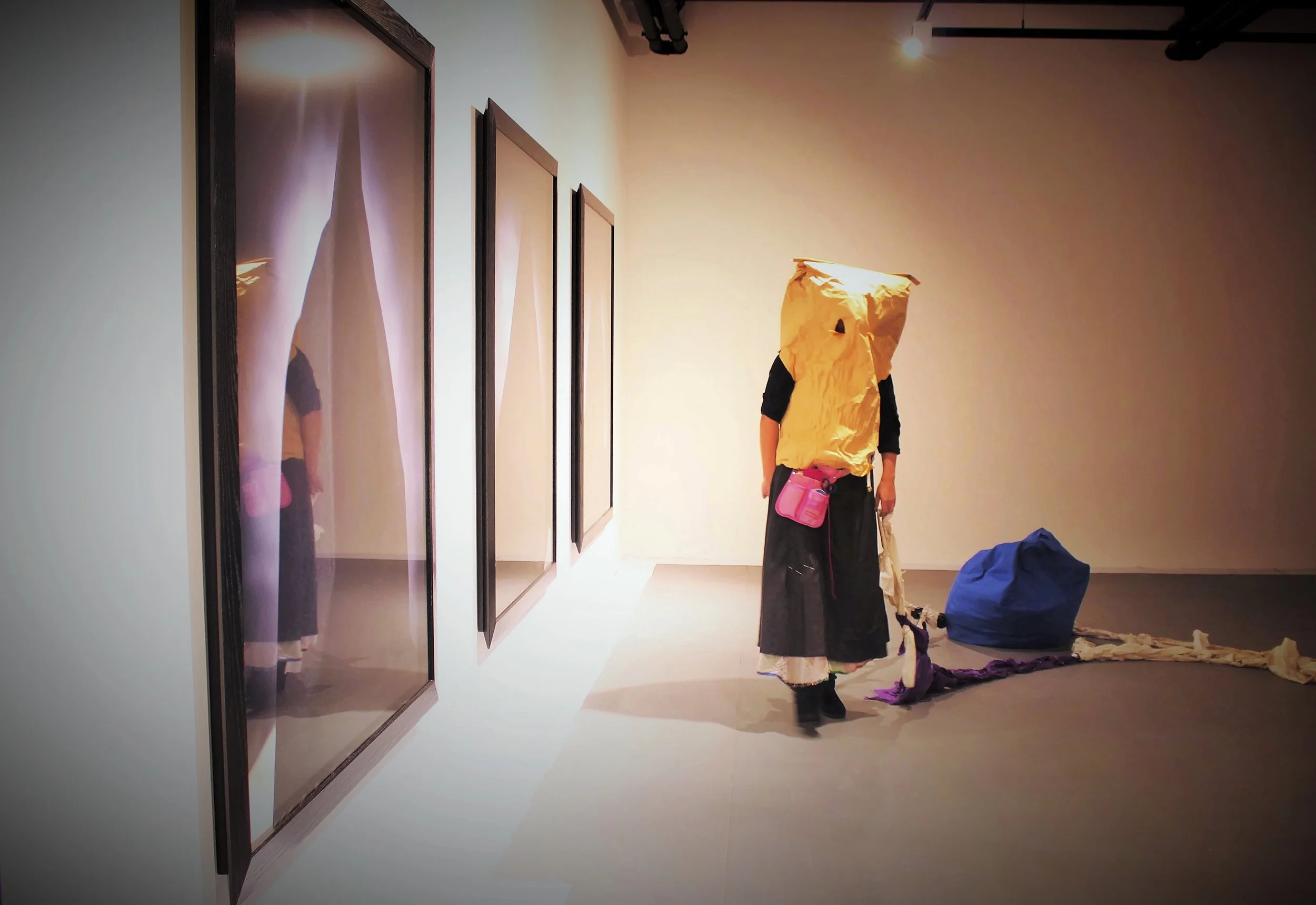 An art installation features a person wearing a costume resembling a yellow paper bag with a face hole. They are standing in a gallery space with framed artworks on the wall and various colored fabric or clothing on the floor.