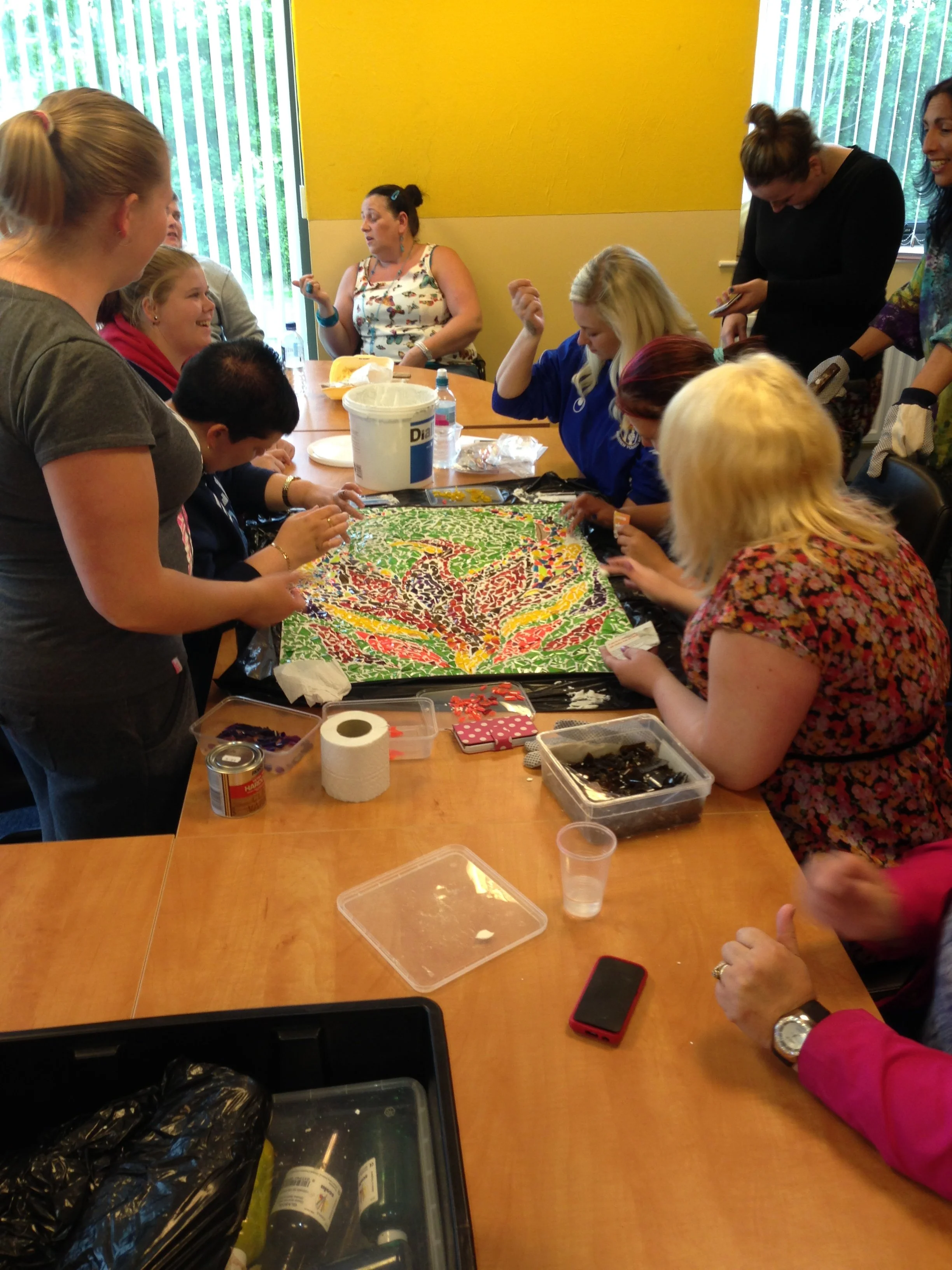 A group of people gathered around a table working on a colorful mosaic or craft project, with various supplies and tools on the table.