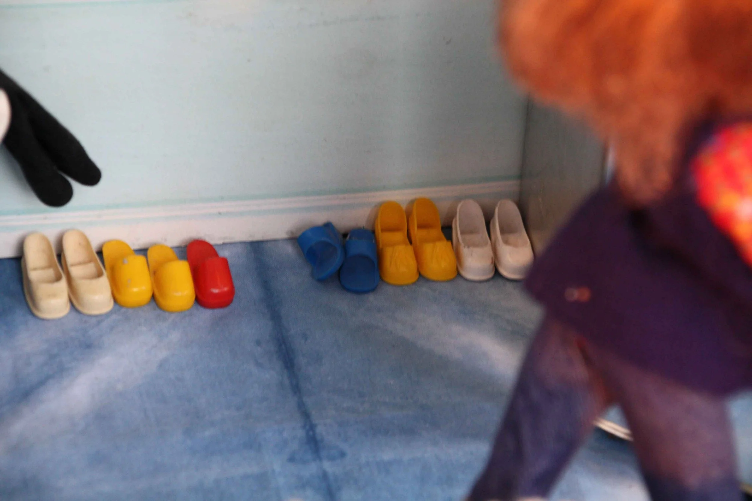 A row of small plastic shoes in various colors including beige, yellow, red, blue, and white, lined up against a wall on a blue floor, with a person partially visible on the right side of the image.