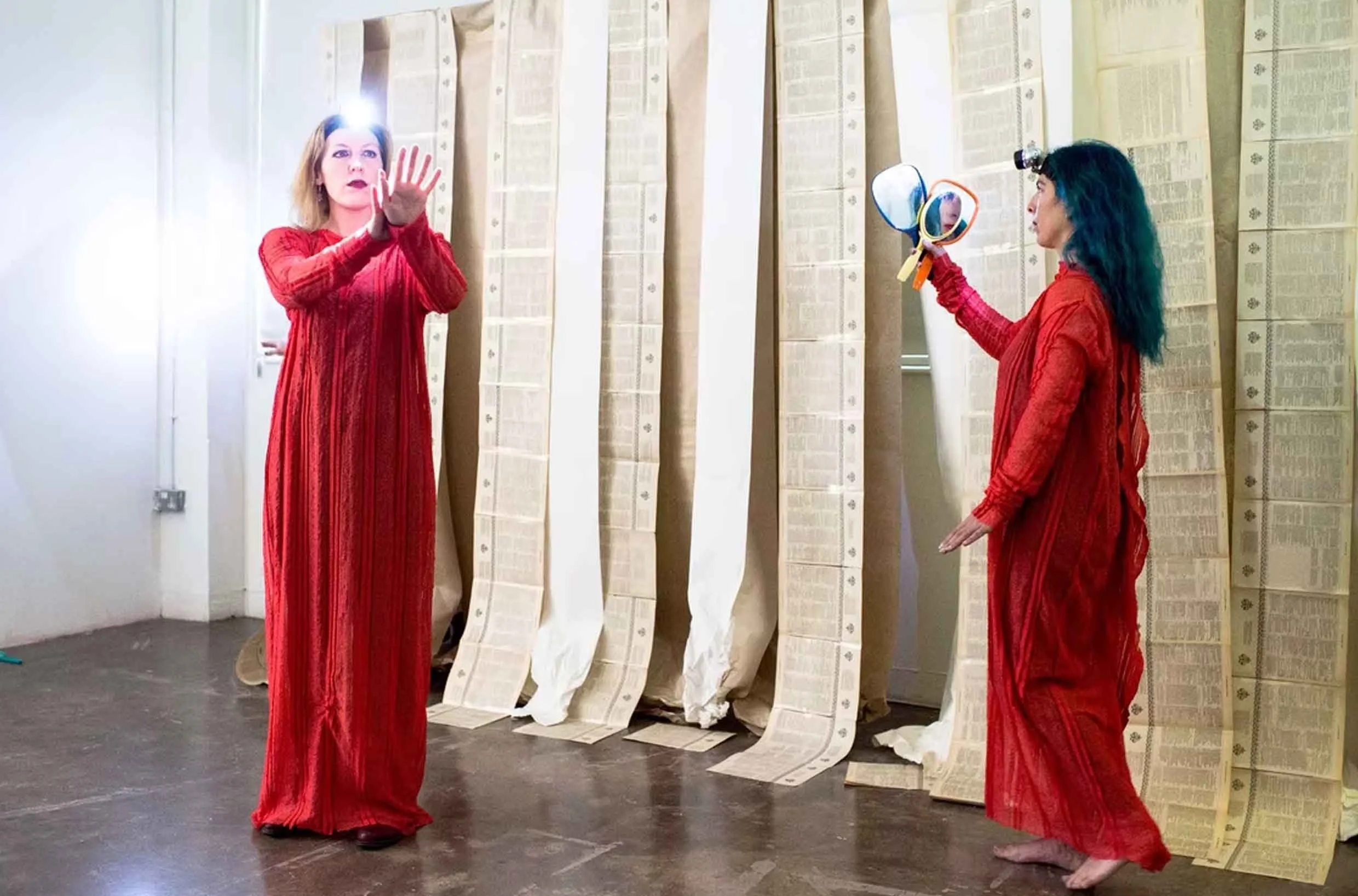 Two women in red dresses stand among long hanging sheets of paper with printed text, one woman facing forward with her hand raised and the other holding a mirror with multiple lenses, in an art installation setting.