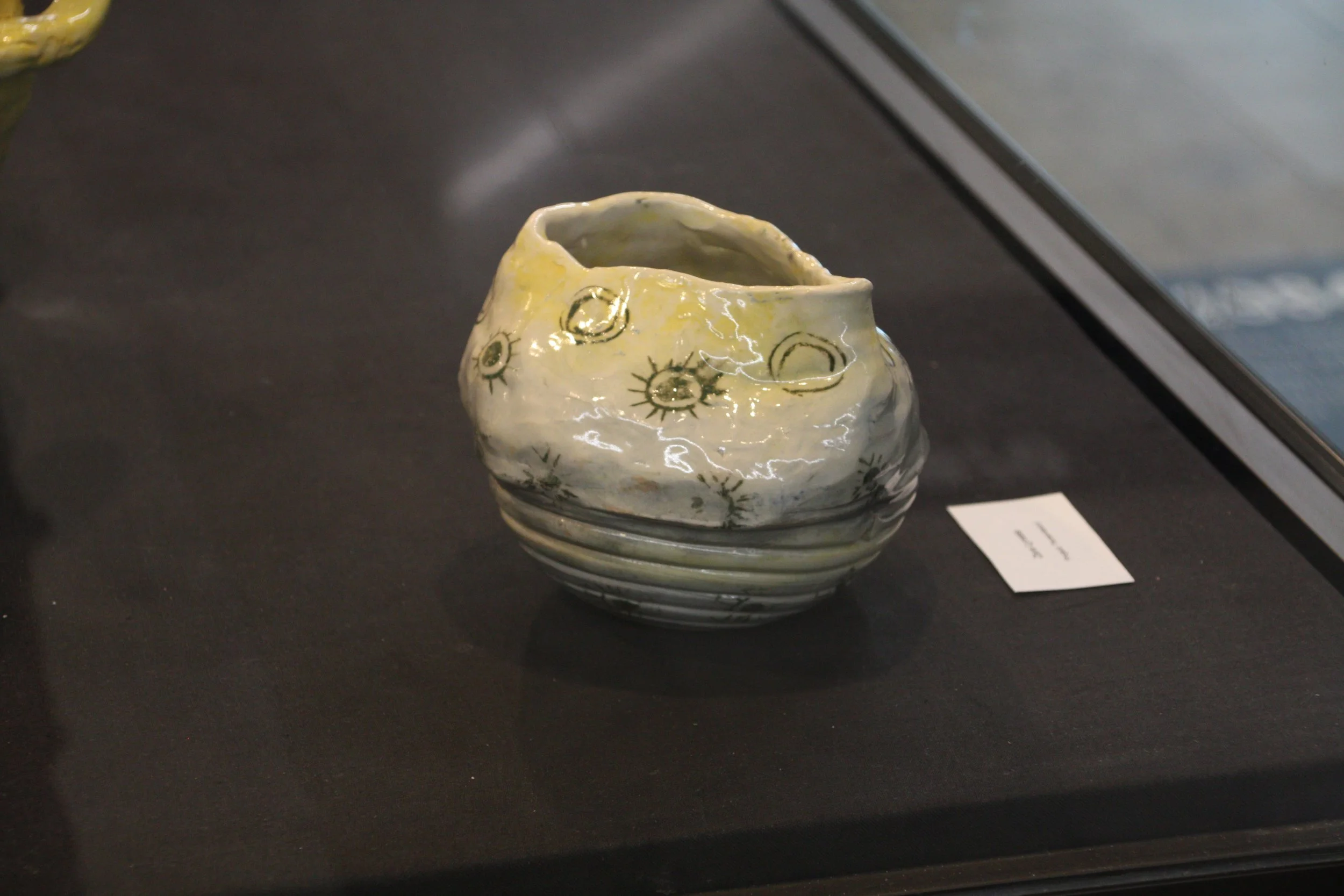 White ceramic bowl with black and yellow sun and moon designs on the surface, displayed on a black surface in a museum or exhibition setting.