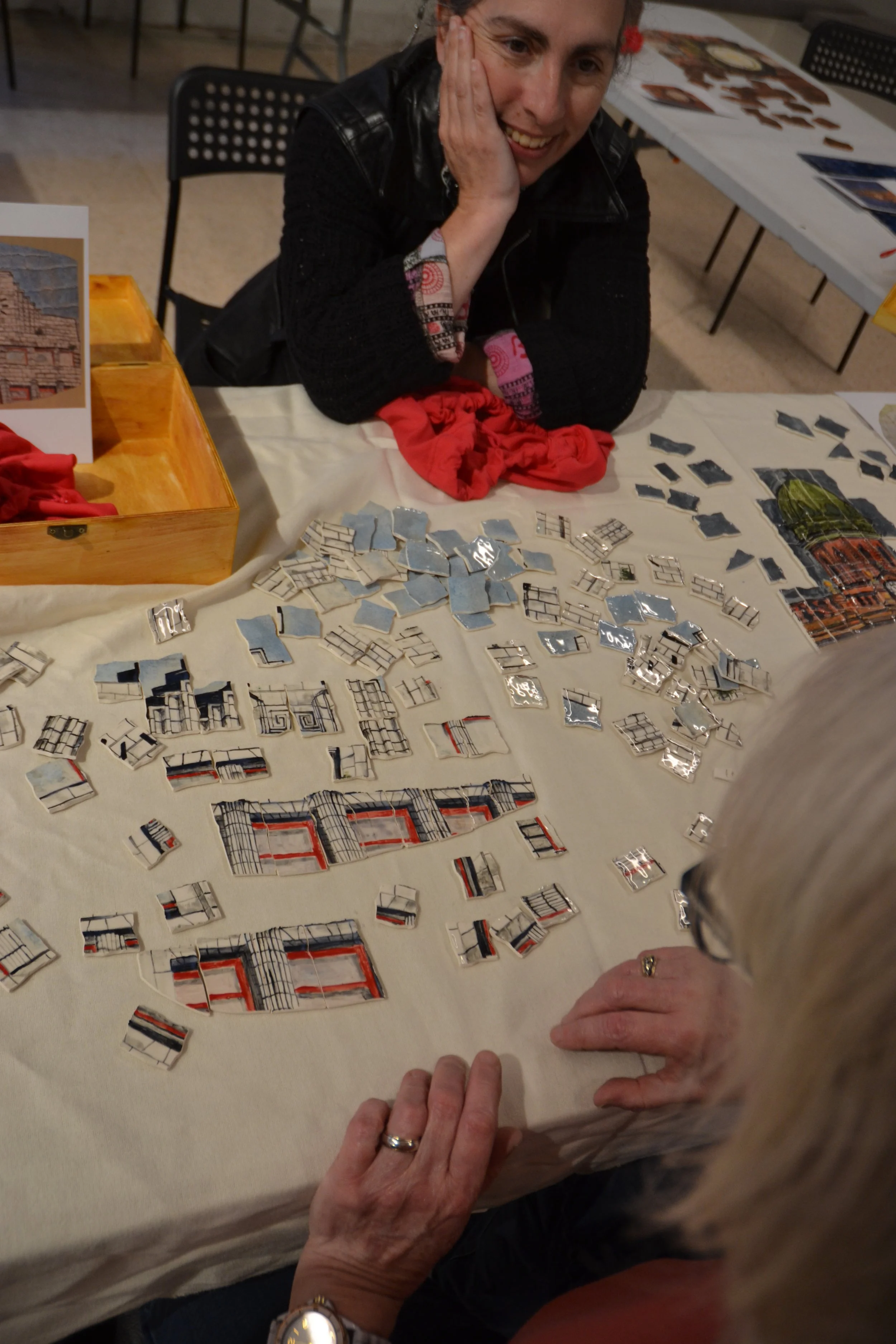 A woman smiling with her hand on her face sitting at a table with a display of small mirror pieces and photographs of buildings, some sketches on paper, and a wooden box with red fabric inside.