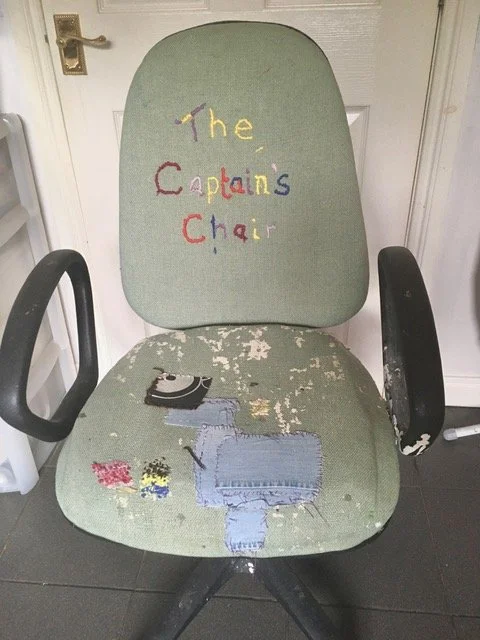 The captain's chair, suburban office or waiting room style, with frayed and torn upholstery and embroidered text reading "The Captains Chair" on the backrest.