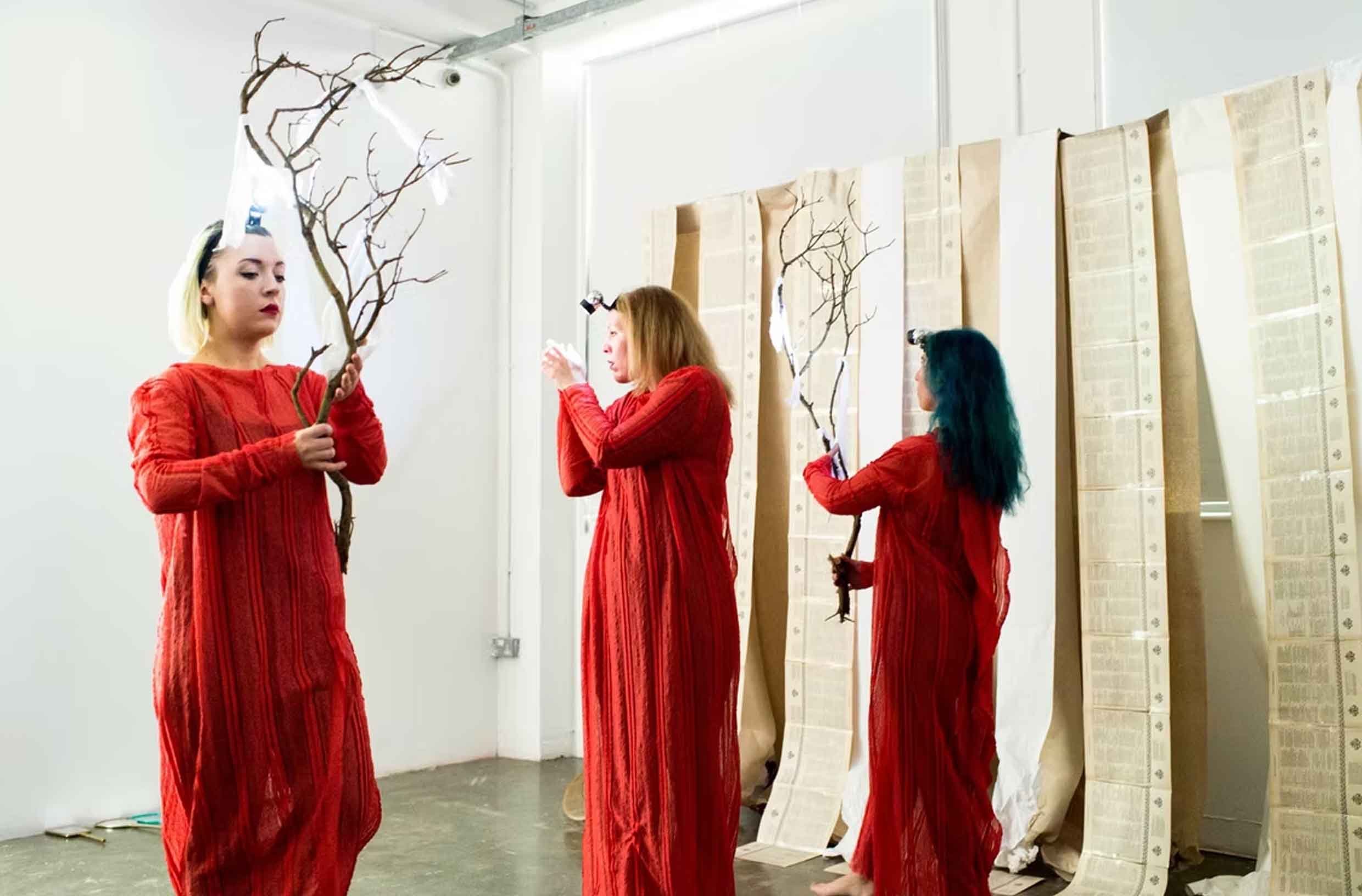 Three women in red dresses holding branches in an art gallery with scrolls or paintings hanging on the wall behind them.