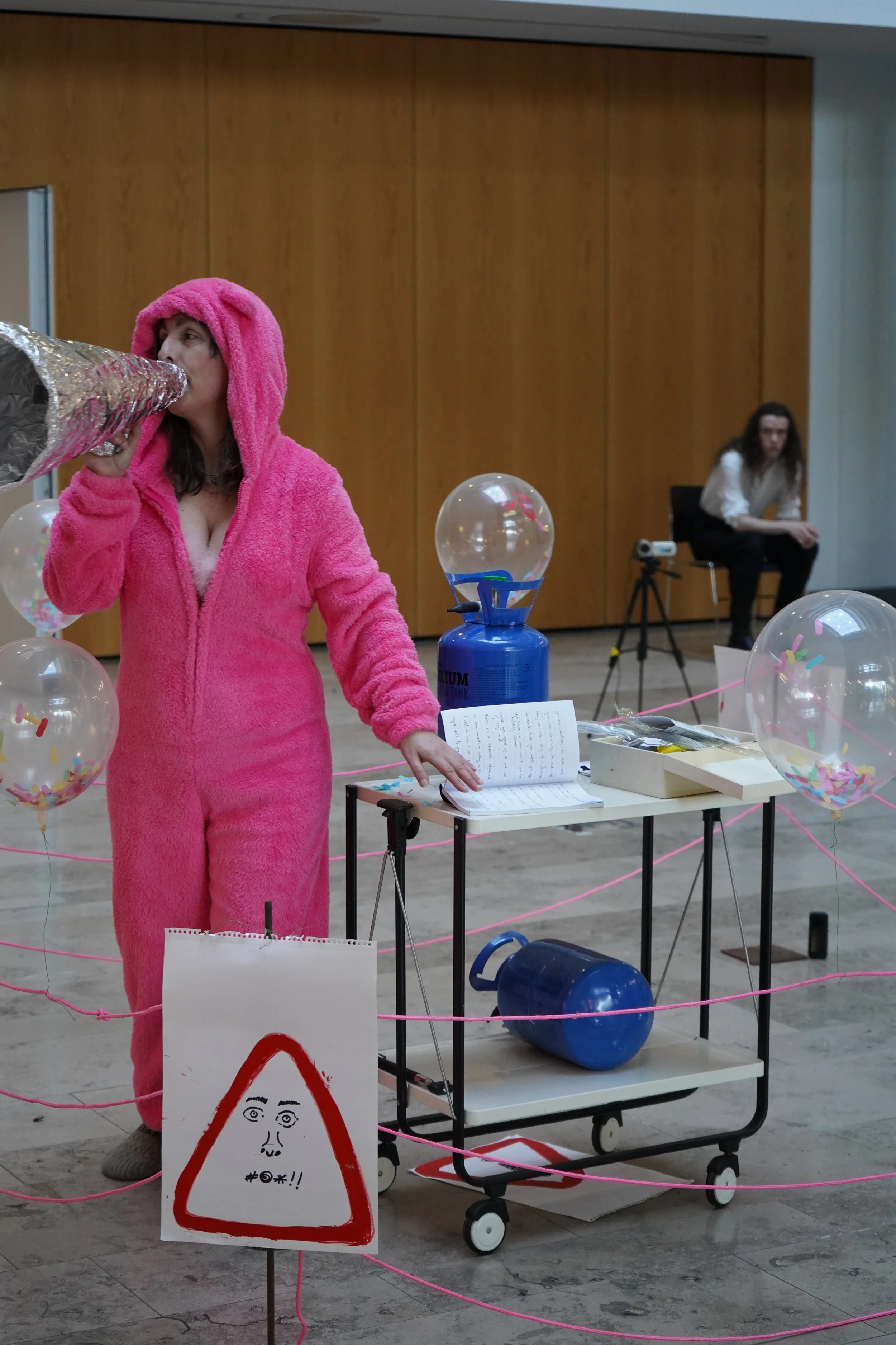 Person in a pink fuzzy onesie blowing up a silver balloon in a room with balloons and signs, with another person sitting in the background.