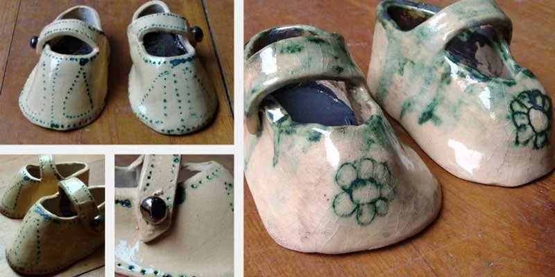 Pair of handmade ceramic baby shoes painted with floral patterns, placed on a wooden surface, next to a pair of painted clay baby shoes with blue and white designs.