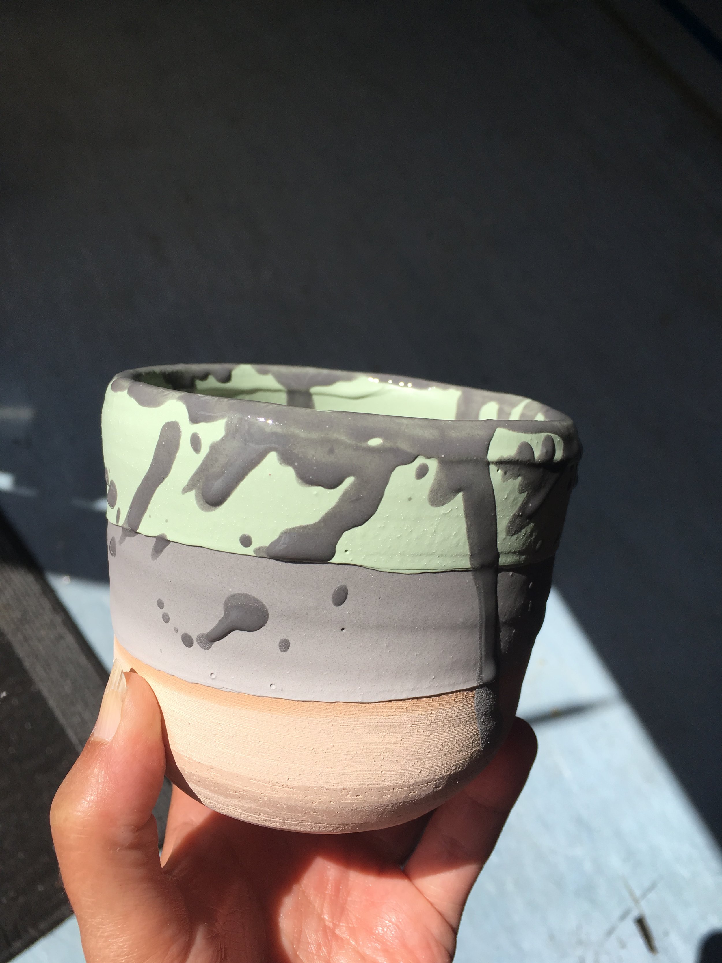 A person's hand holding a ceramic cup with gray and white drip glaze design, set against a dark background.