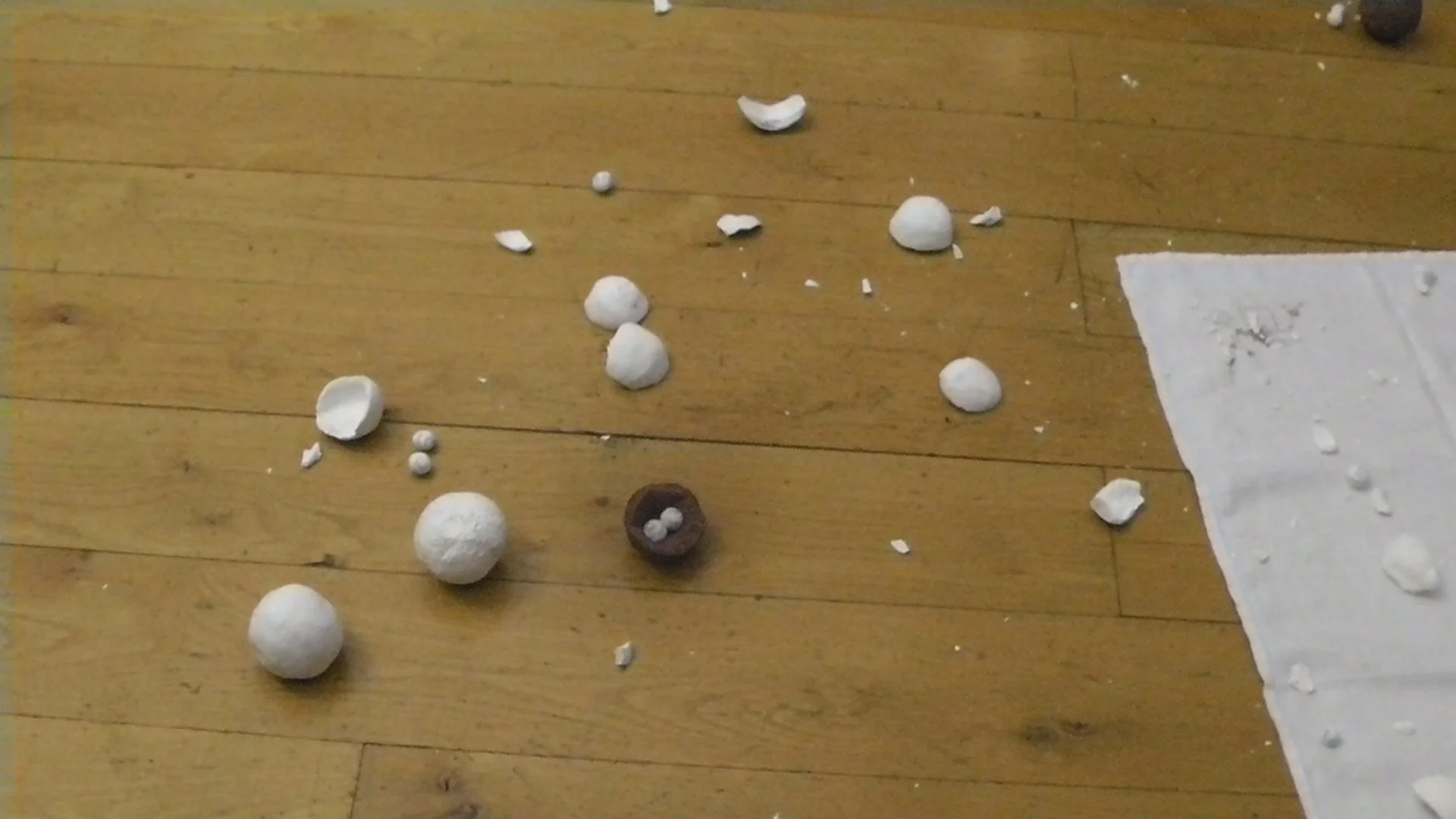 Broken ceramic balls and fragments scattered on a wooden floor with a paper towel to the right.