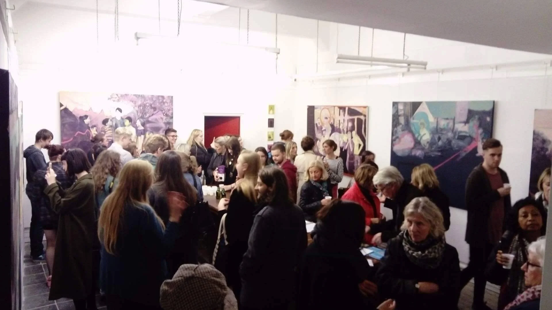 A crowded art exhibition with people viewing and socializing in an indoor gallery space, featuring large colorful paintings on the white walls.