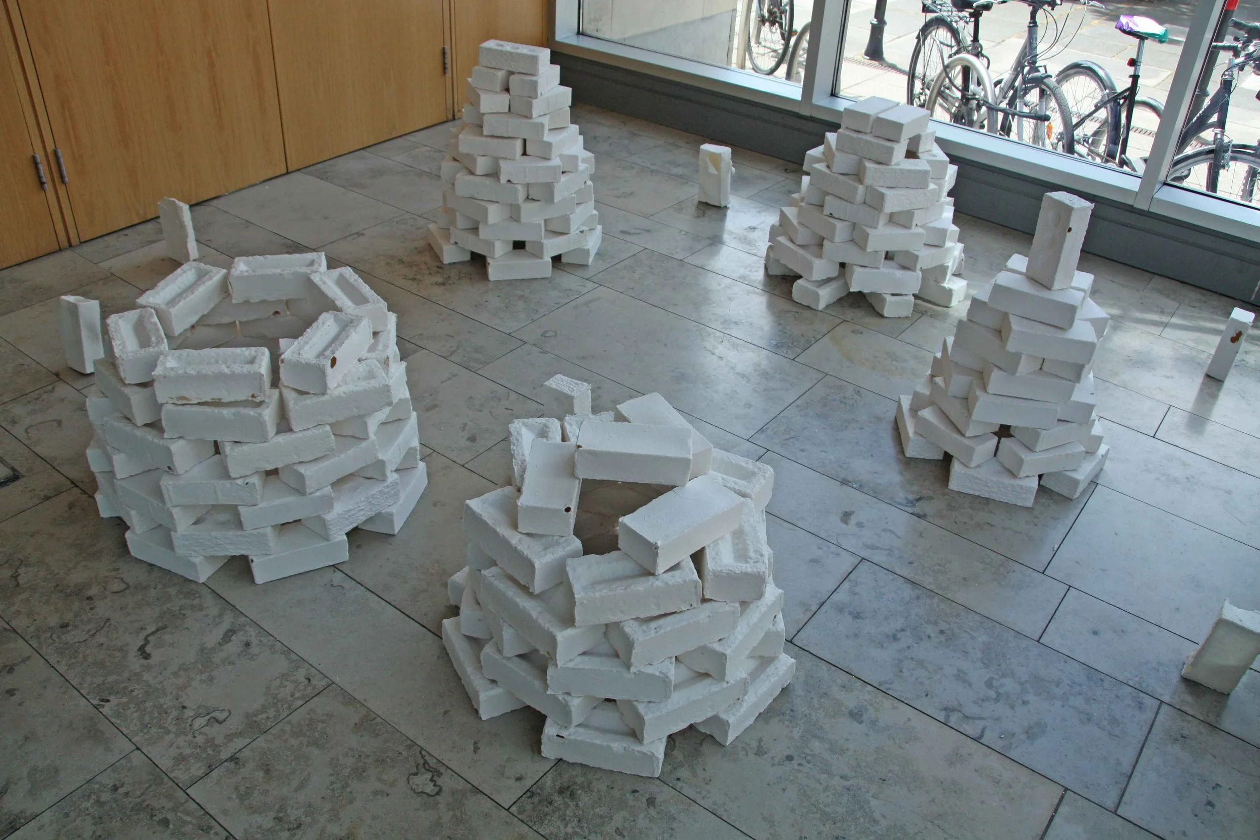 Indoor scene with multiple stacks of white ceramic arranged in round chimney shapes on a tiled floor near a glass window, with bicycles visible outside.