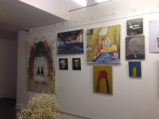 Gallery wall with multiple colorful paintings, a floral cage-like art piece with flowers, and a cluster of white flowers in the foreground.