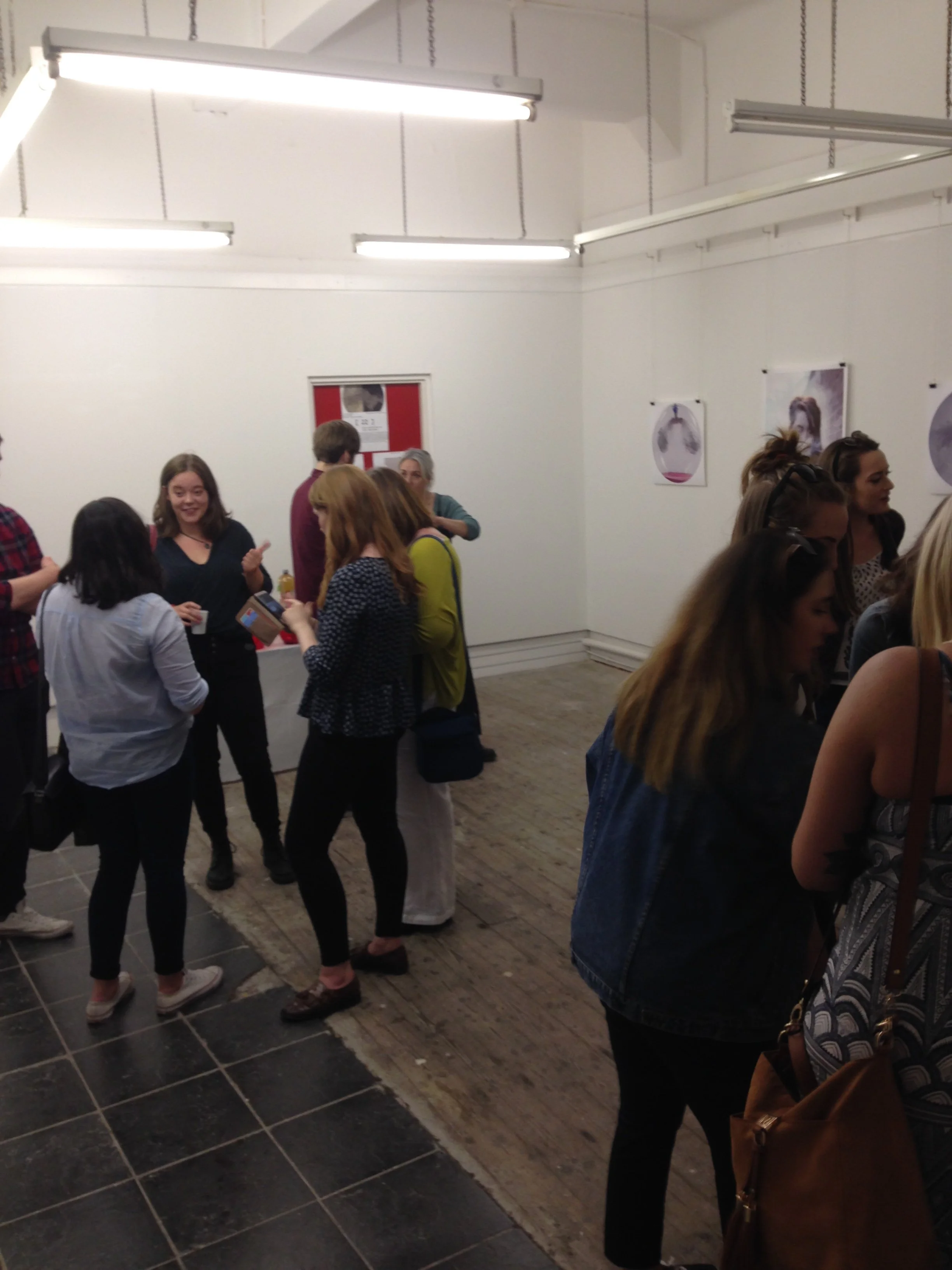 People gathered in an art gallery, engaged in conversations, with white walls and artwork displayed, under ceiling lights.