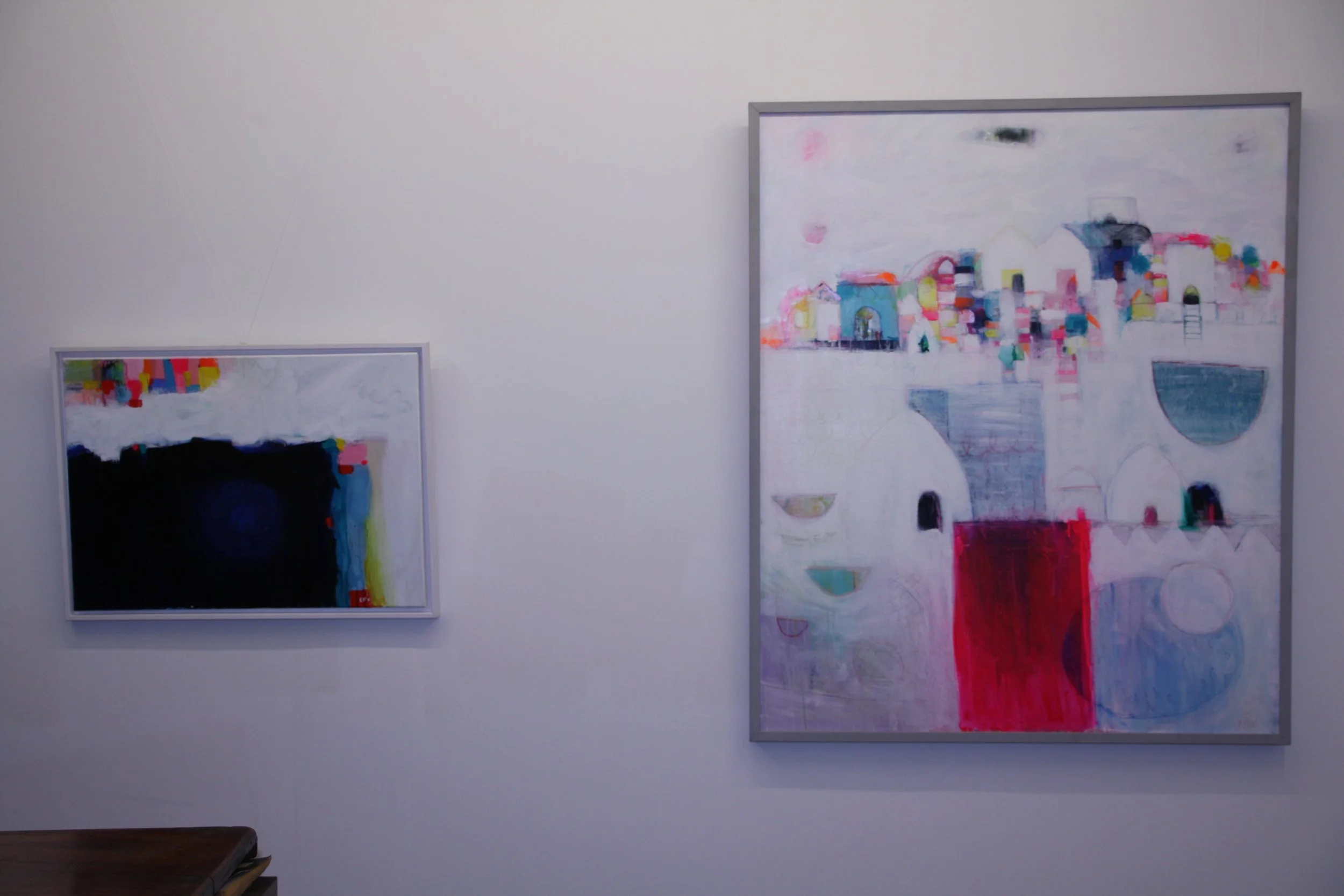 Two abstract paintings hanging on a white gallery wall, one smaller with darker colors and the other larger with bright colors and geometric shapes.