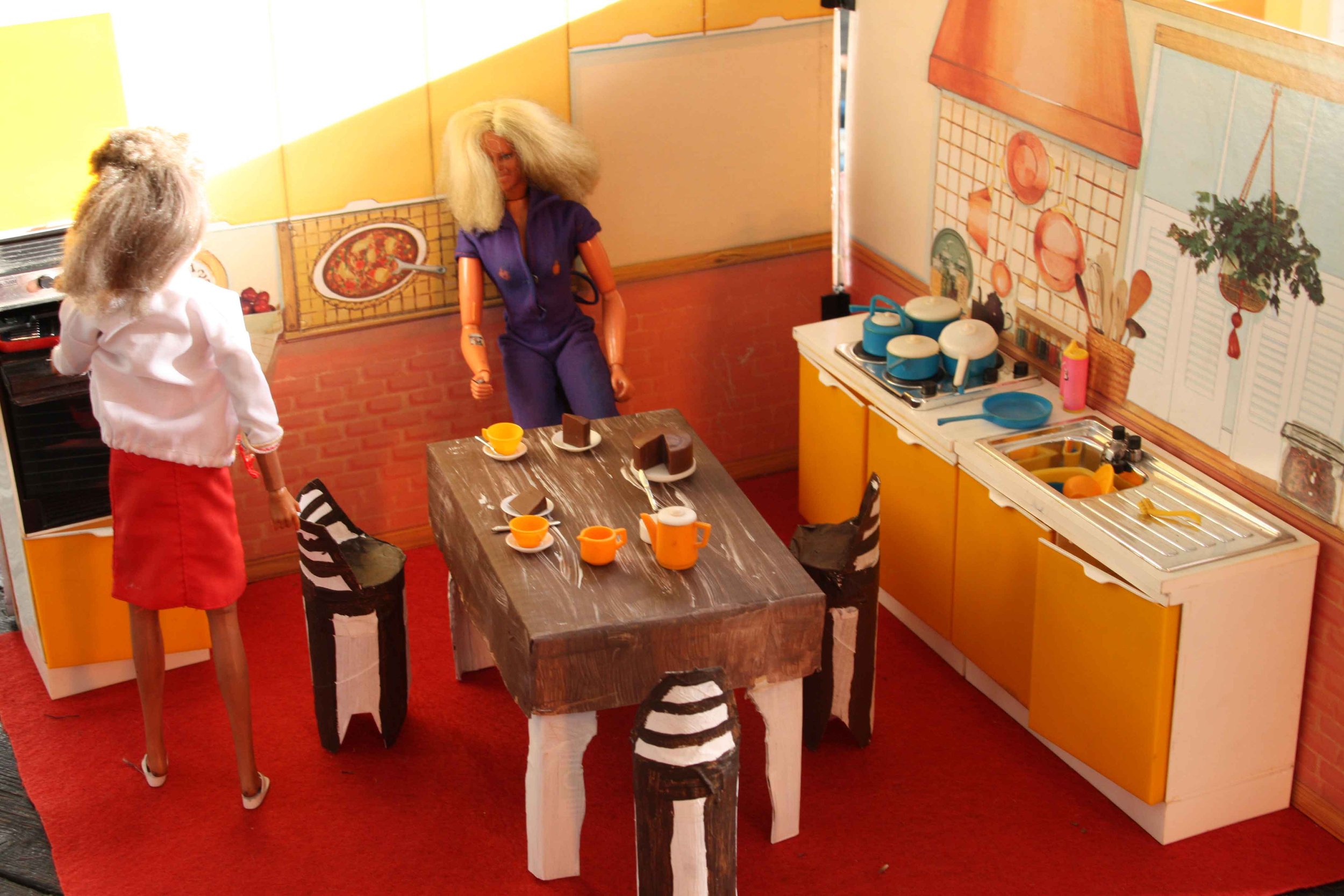 Dollhouse kitchen scene with yellow cabinets, a small wooden dining table set with colorful cups and plates, a faux brick wall, and decorative wall art.