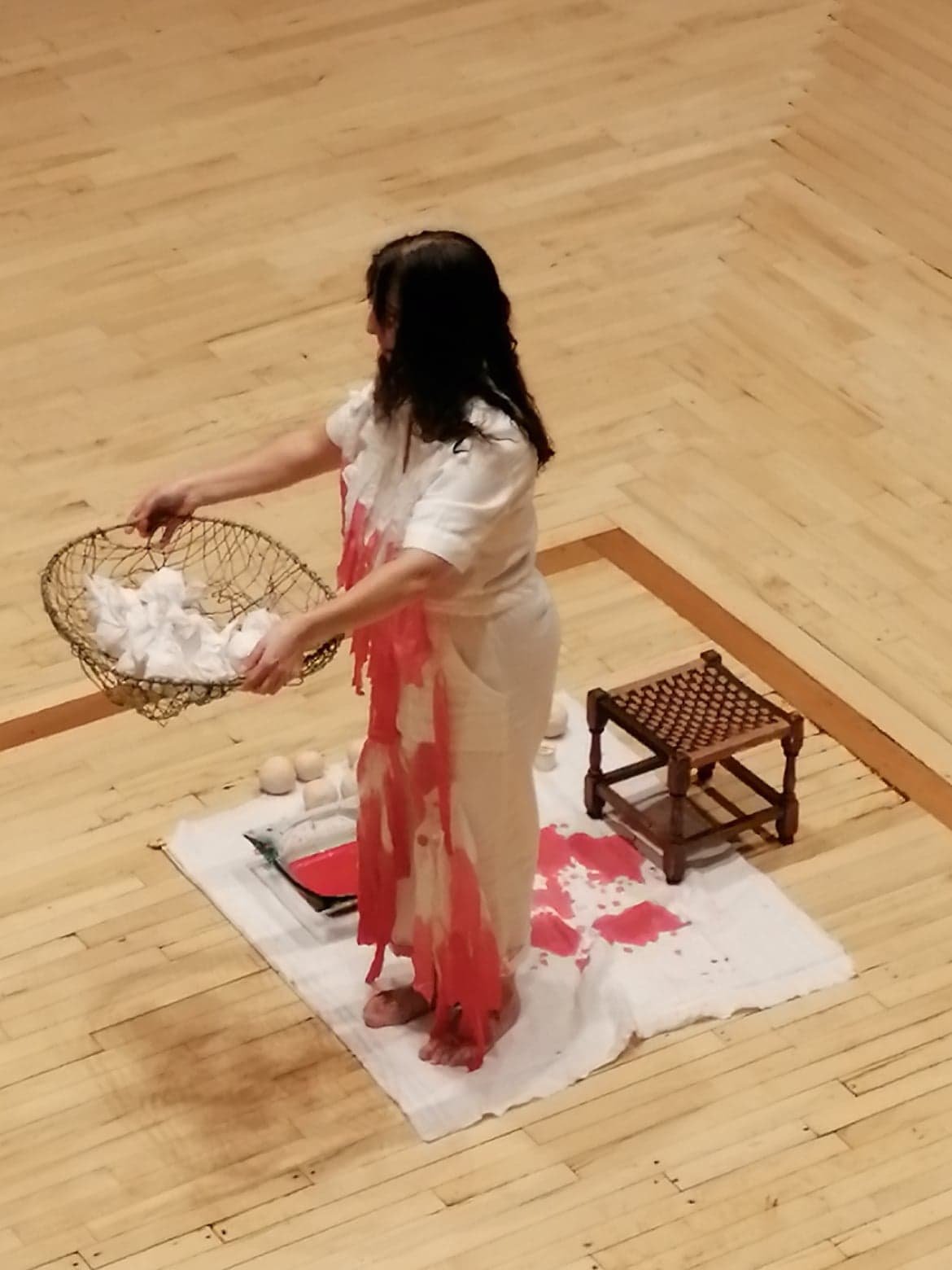 A woman dressed in white and red is standing on a white cloth stained with red, holding a basket filled with white cloths or tissues, with a small wooden stool nearby on a wooden floor.
