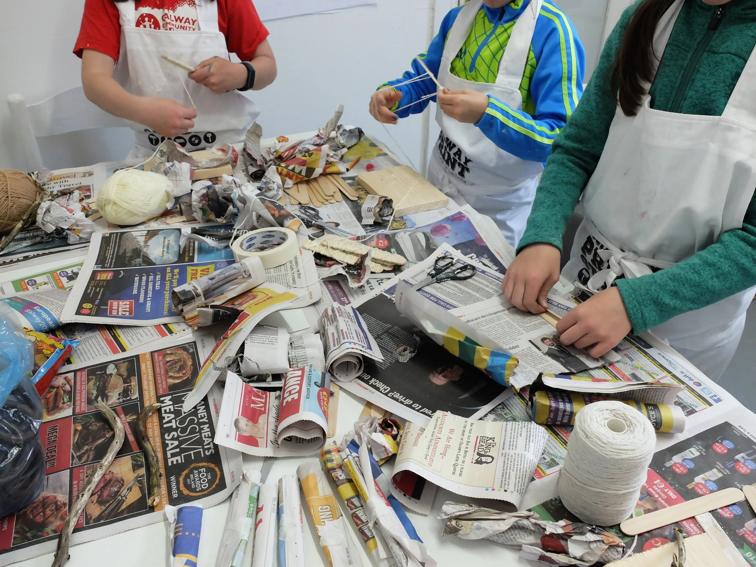 Children wearing aprons working on a craft project at a table covered with crumpled newspapers, yarn, and wooden craft sticks.