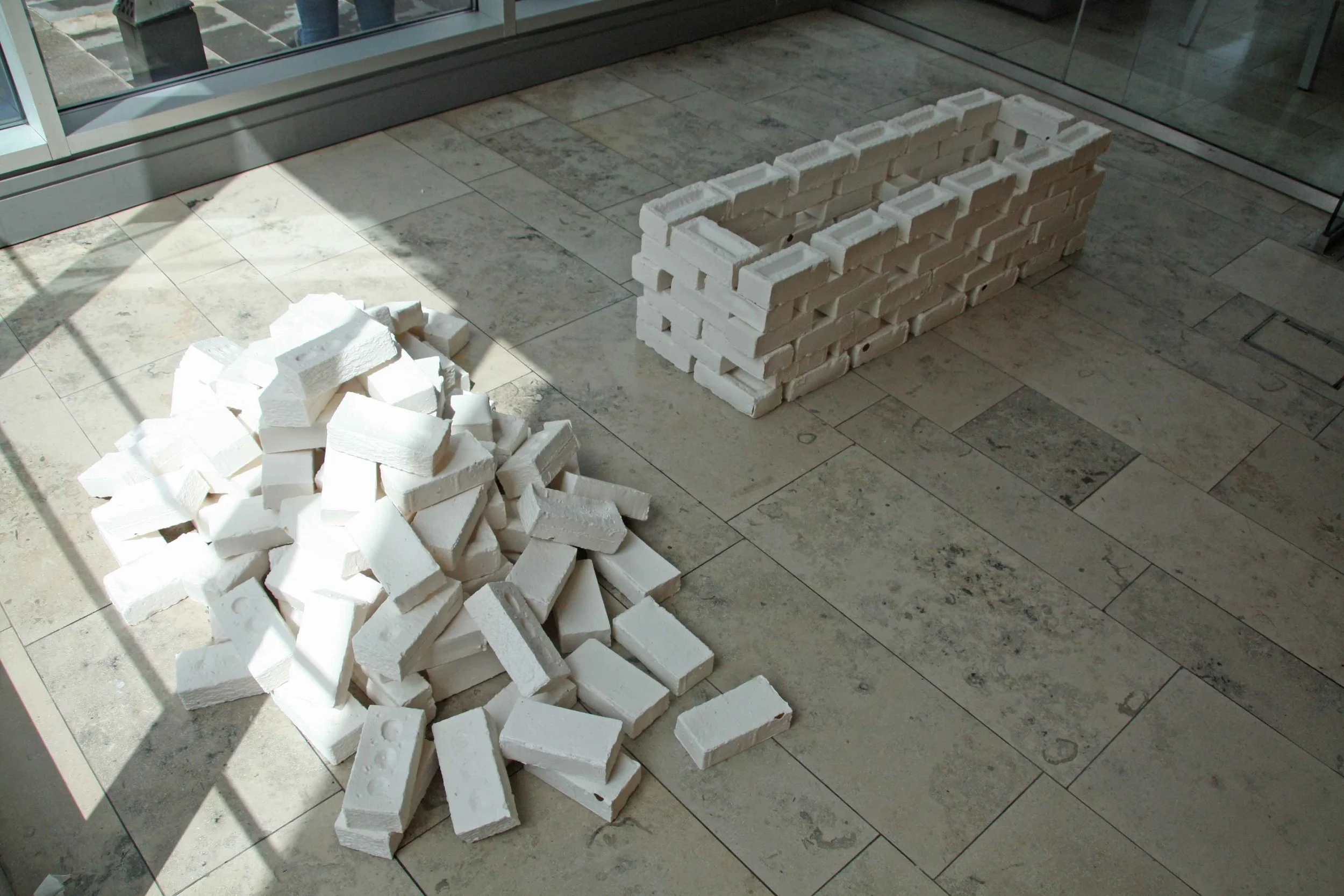 A pile of white ceramic bricks and a stack of ceramic  bricks arranged in a rectangular shape on a tiled floor next to large glass windows.