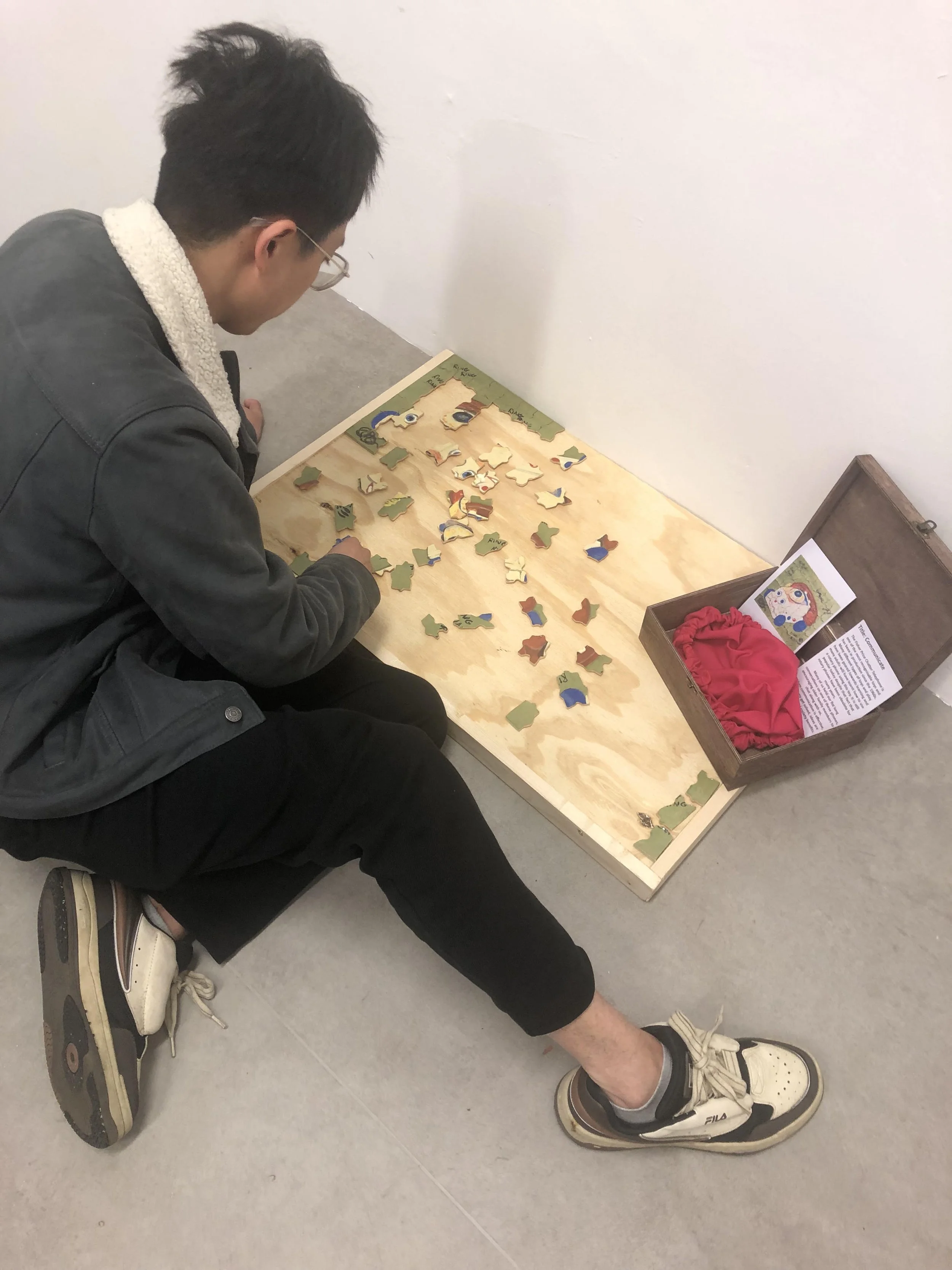 A person wearing glasses and a black jacket with a fleece collar is crouching down on the floor, working on a puzzle on a wooden table. The puzzle pieces are scattered across the table. There is a wooden box on the right side of the table with some p