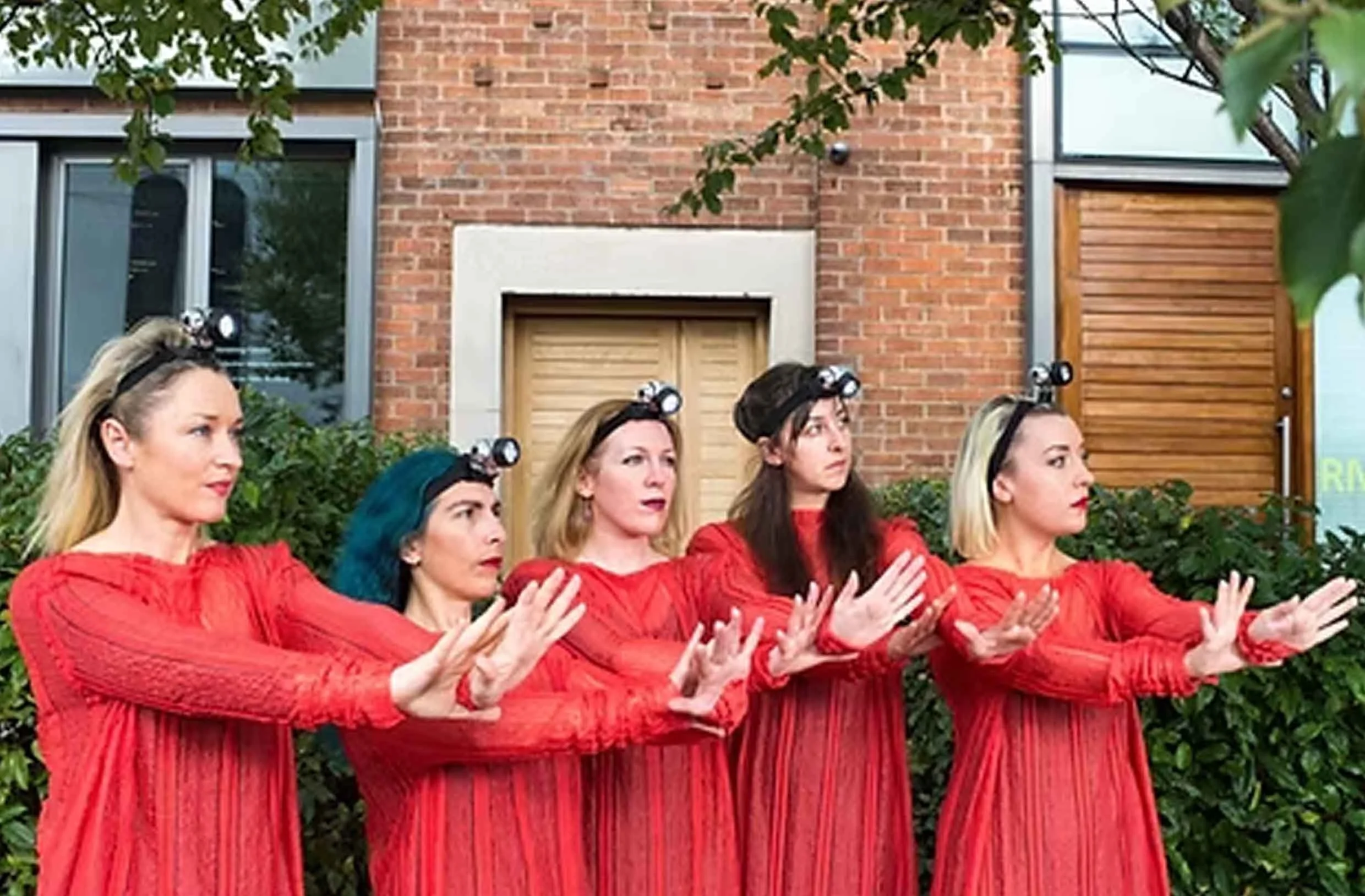 Group of women wearing red dresses, with headlamps, performing a synchronized dance outdoors in front of a brick building and trees.