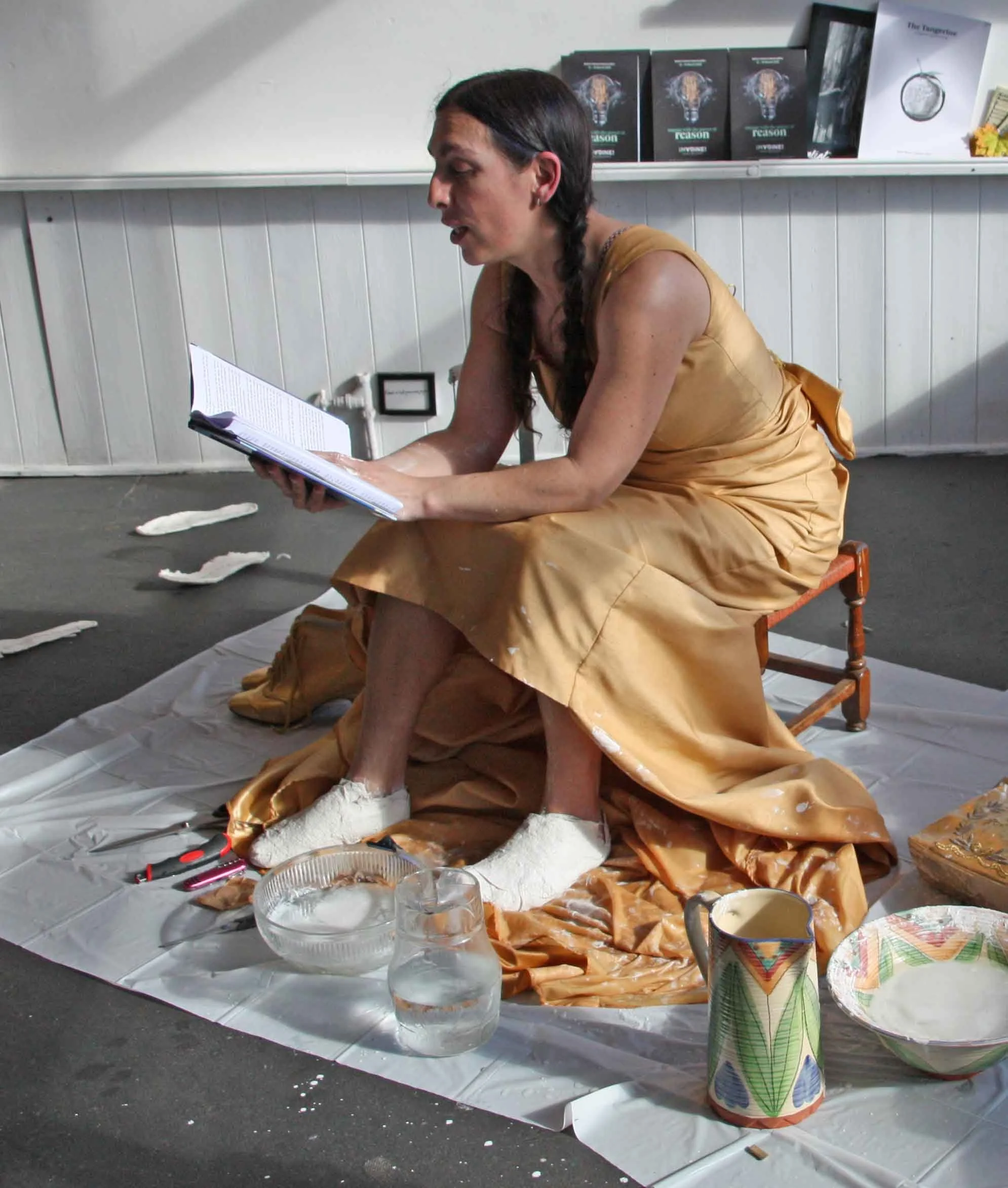 A woman dressed in a golden outfit with a braid in her hair is sitting on a small chair, reading a book, among torn paper and objects on the floor. In front of her, there are bowls of water, a colorful cup, and other items on a white sheet. Behind he