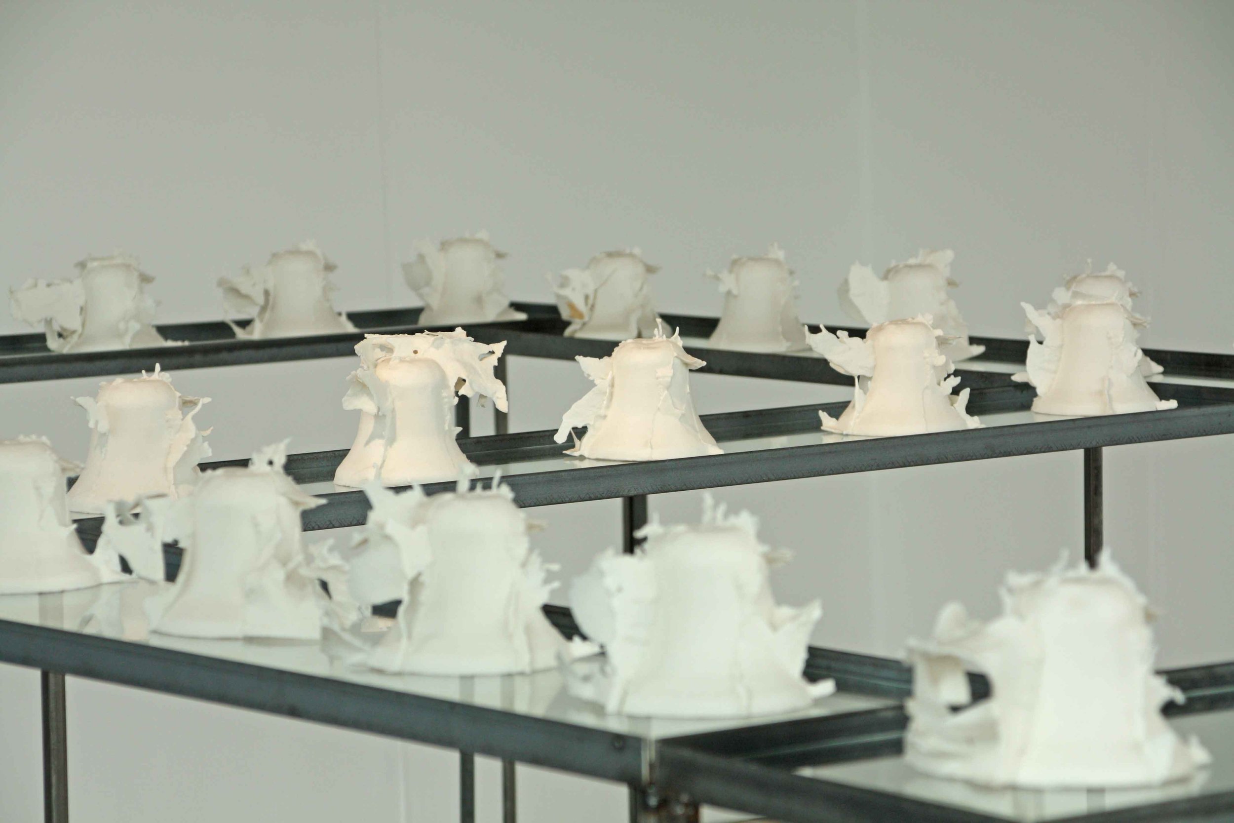 Multiple white ceramic pieces resembling broken or unfinished beels displayed on multi-tiered black shelves in a gallery or exhibit setting.