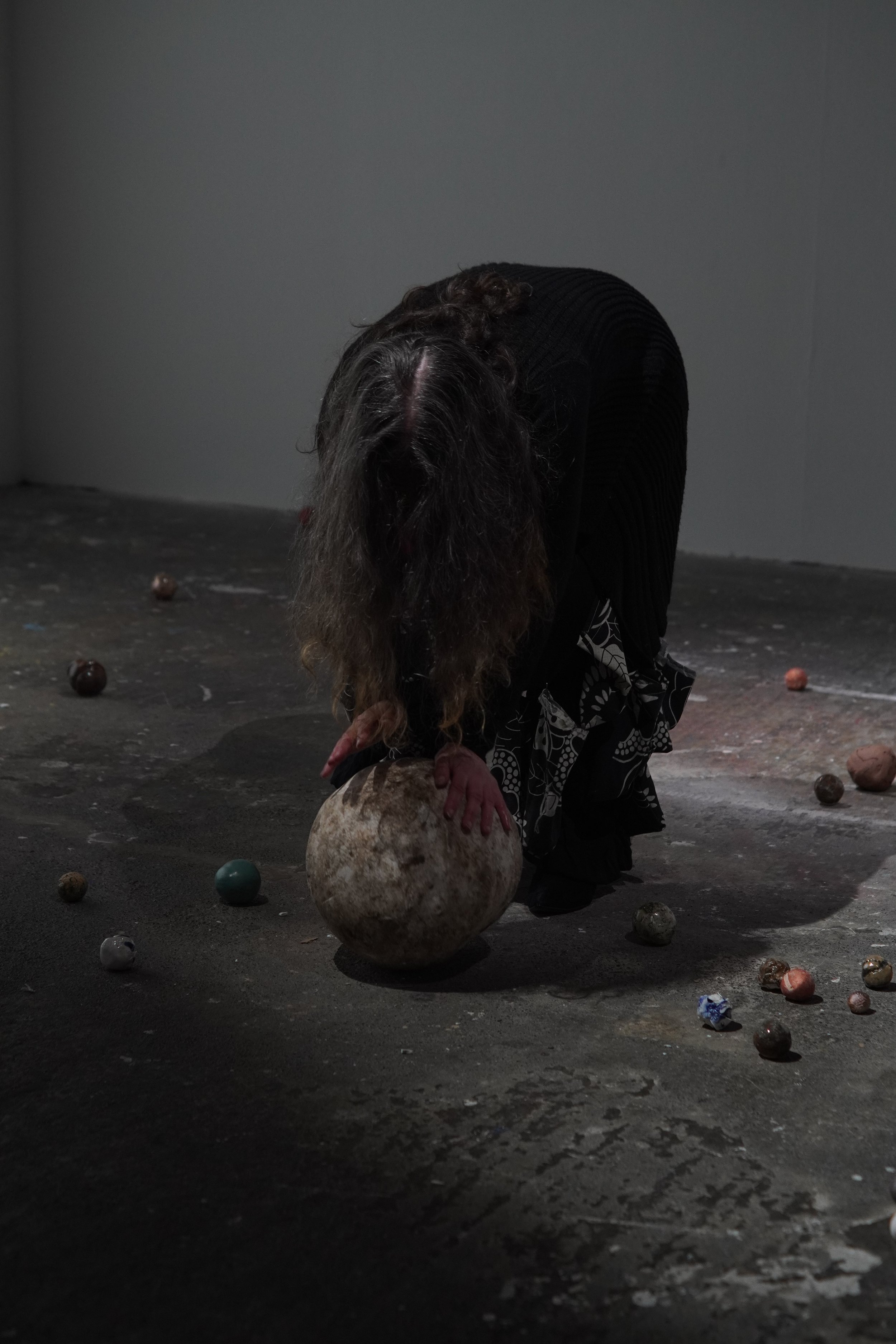 A person with long hair wearing dark clothing, bending down and holding a large round object, surrounded by smaller colorful balls on a gray floor in a minimalistic space.