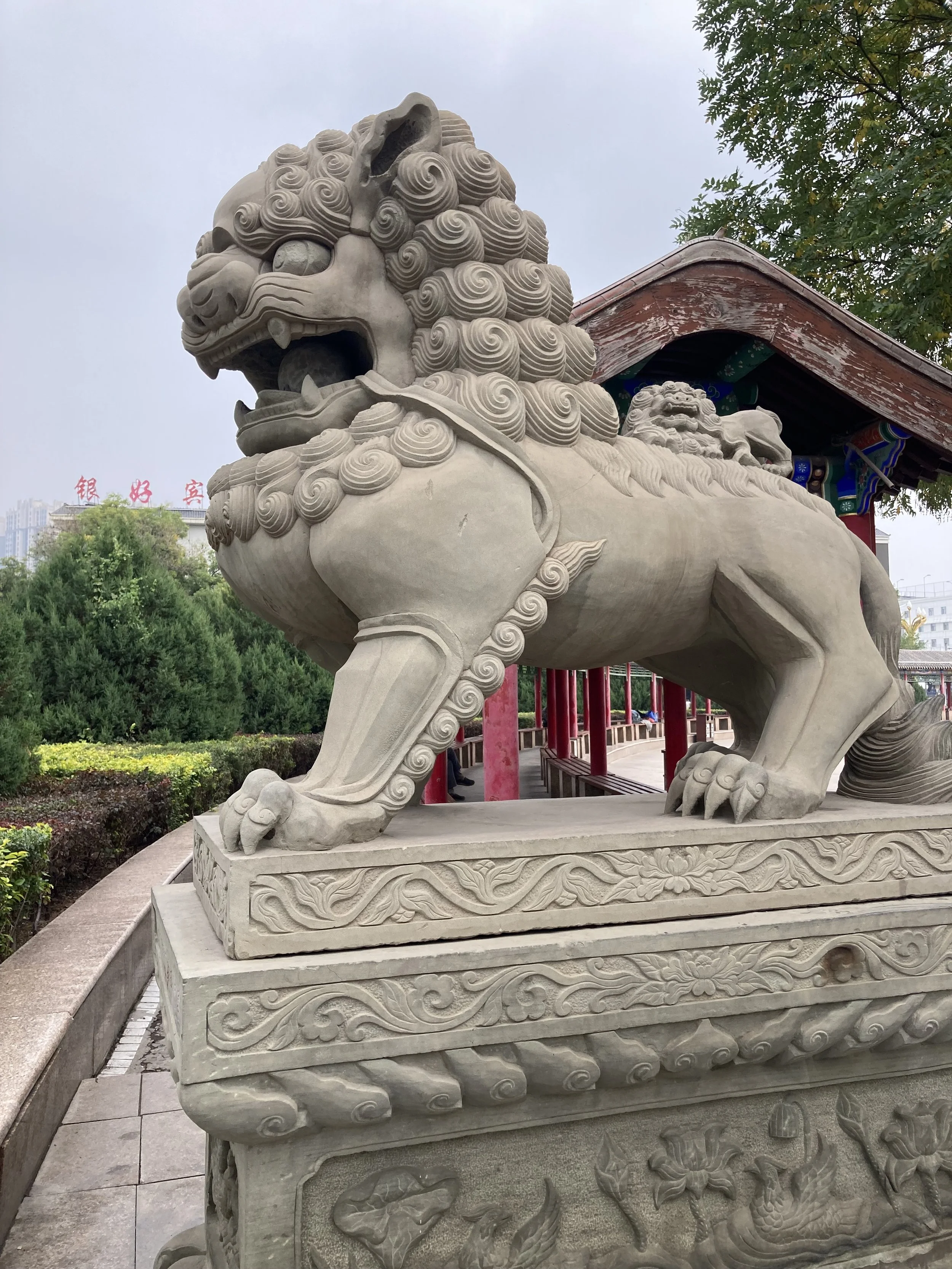 A large stone lion sculpture on a decorated base, with traditional Chinese architectural elements in the background and trees around.