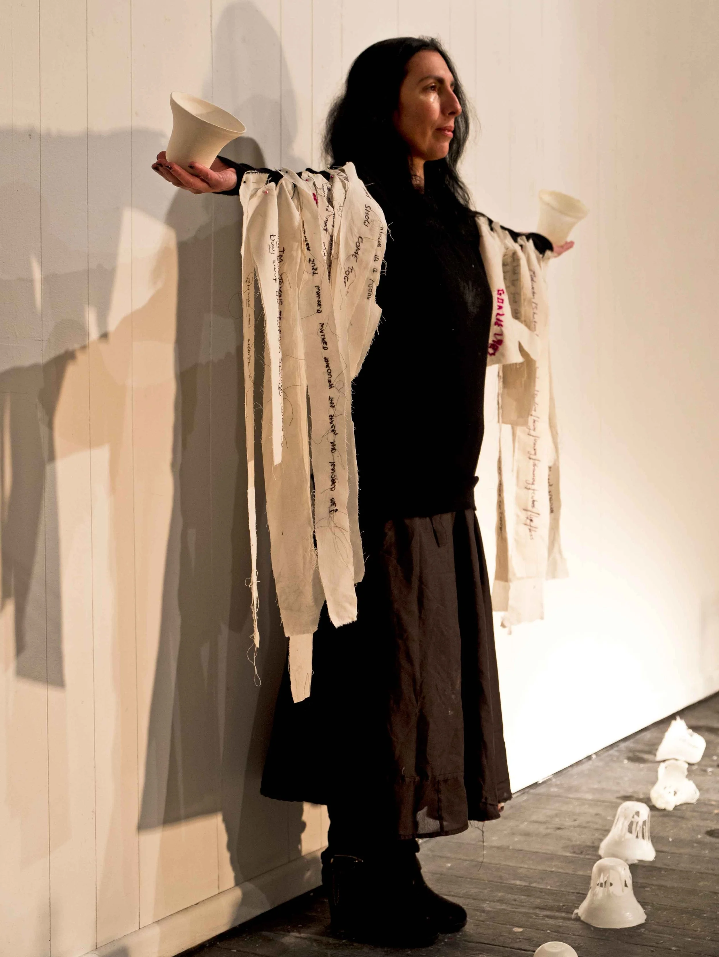 Woman with long dark hair standing with arms outstretched, holding a white cup in each hand, against a light-colored wall, with art installations hanging from her sleeves and objects on the floor.