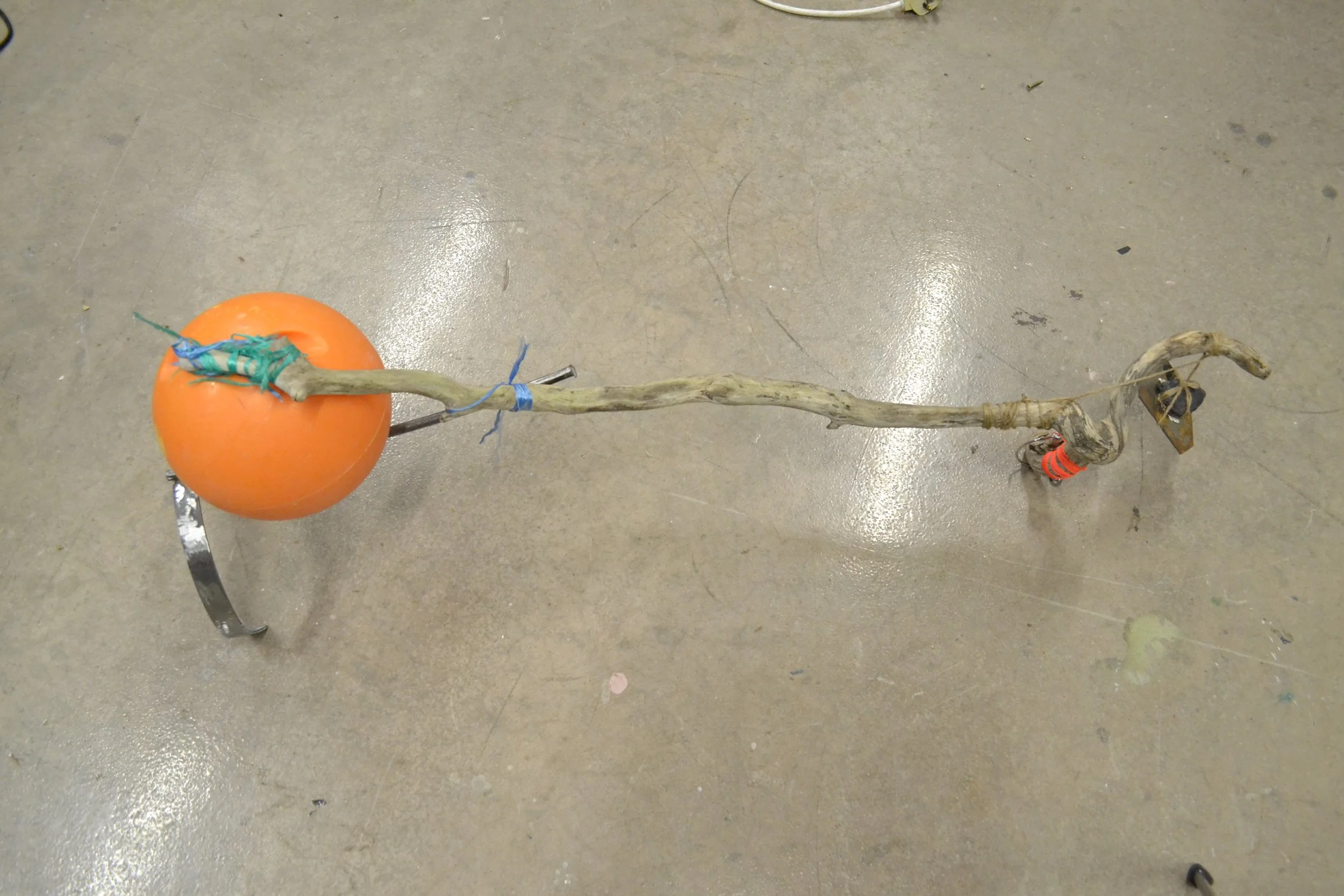 A makeshift walking cane made from a tree branch with a balloon tied around the top and colorful tape wrapped around parts of the branch, resting on a concrete floor.