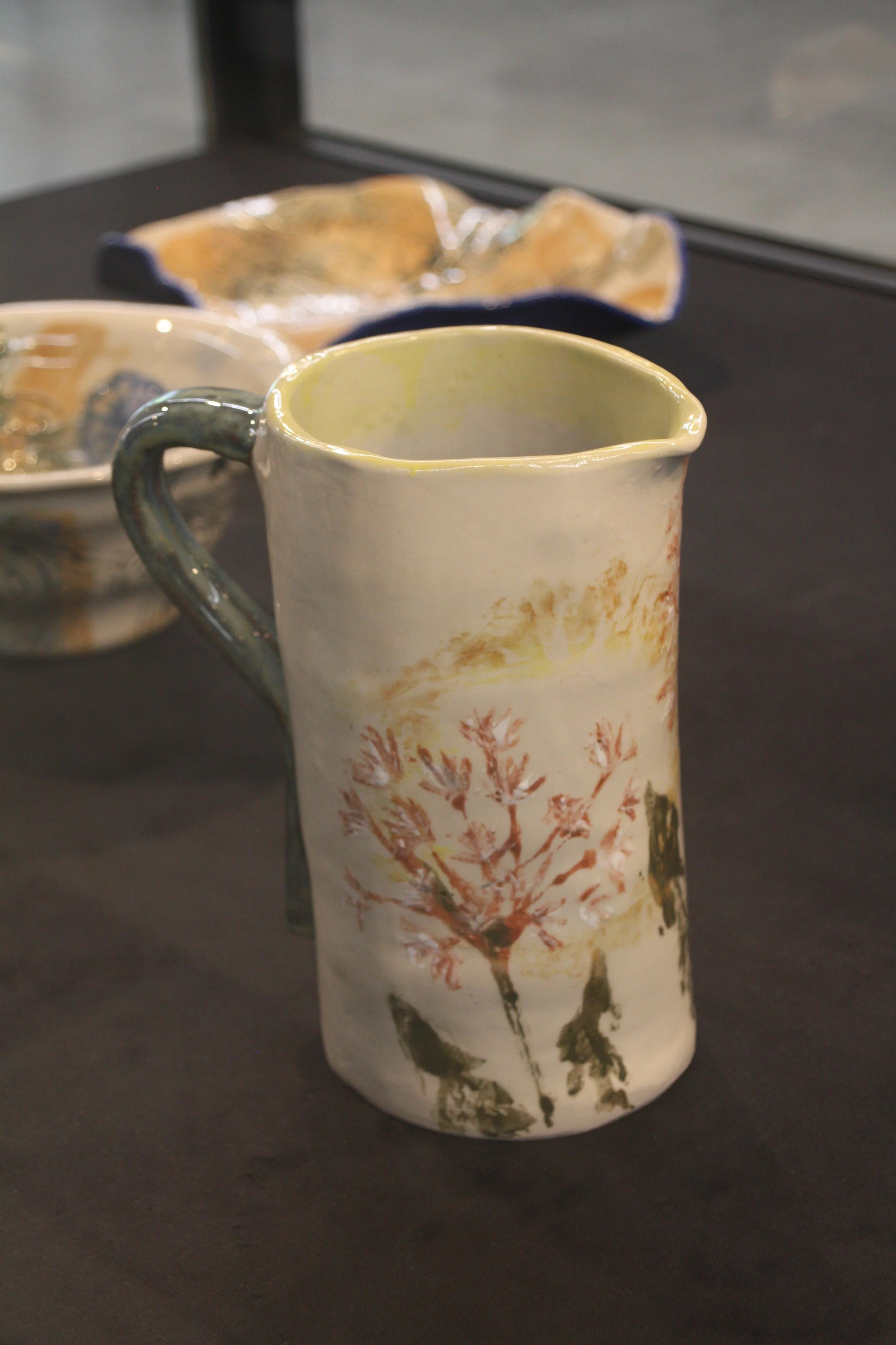 A tall ceramic mug with a tree branch design on a dark surface, with ceramic bowls in the background.