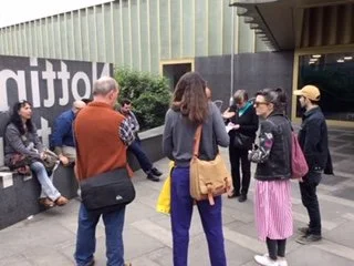 Group of people standing and talking outside a building with a black wall and greenery.