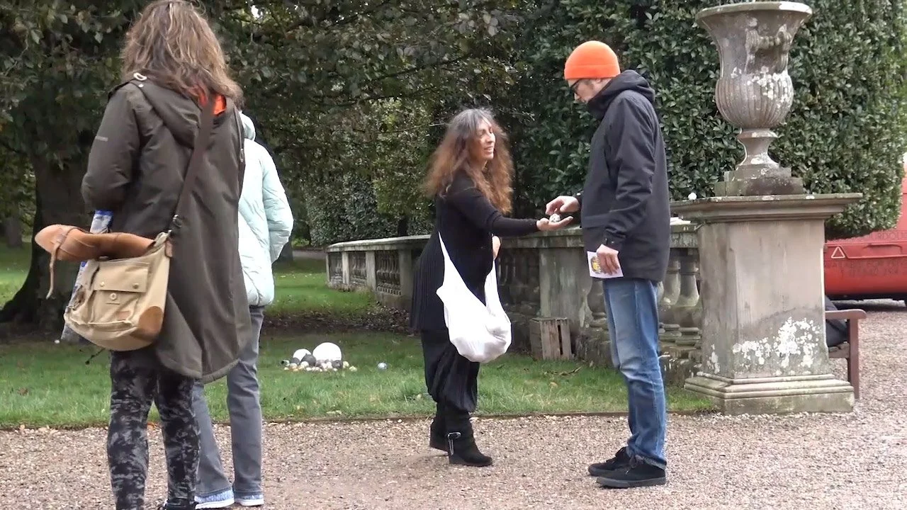 A woman with long curly hair wearing black and white clothing is exchanging something with a man wearing a black jacket and orange beanie. Two additional women stand nearby in a park with greenery, a stone fence, and a large stone planter.
