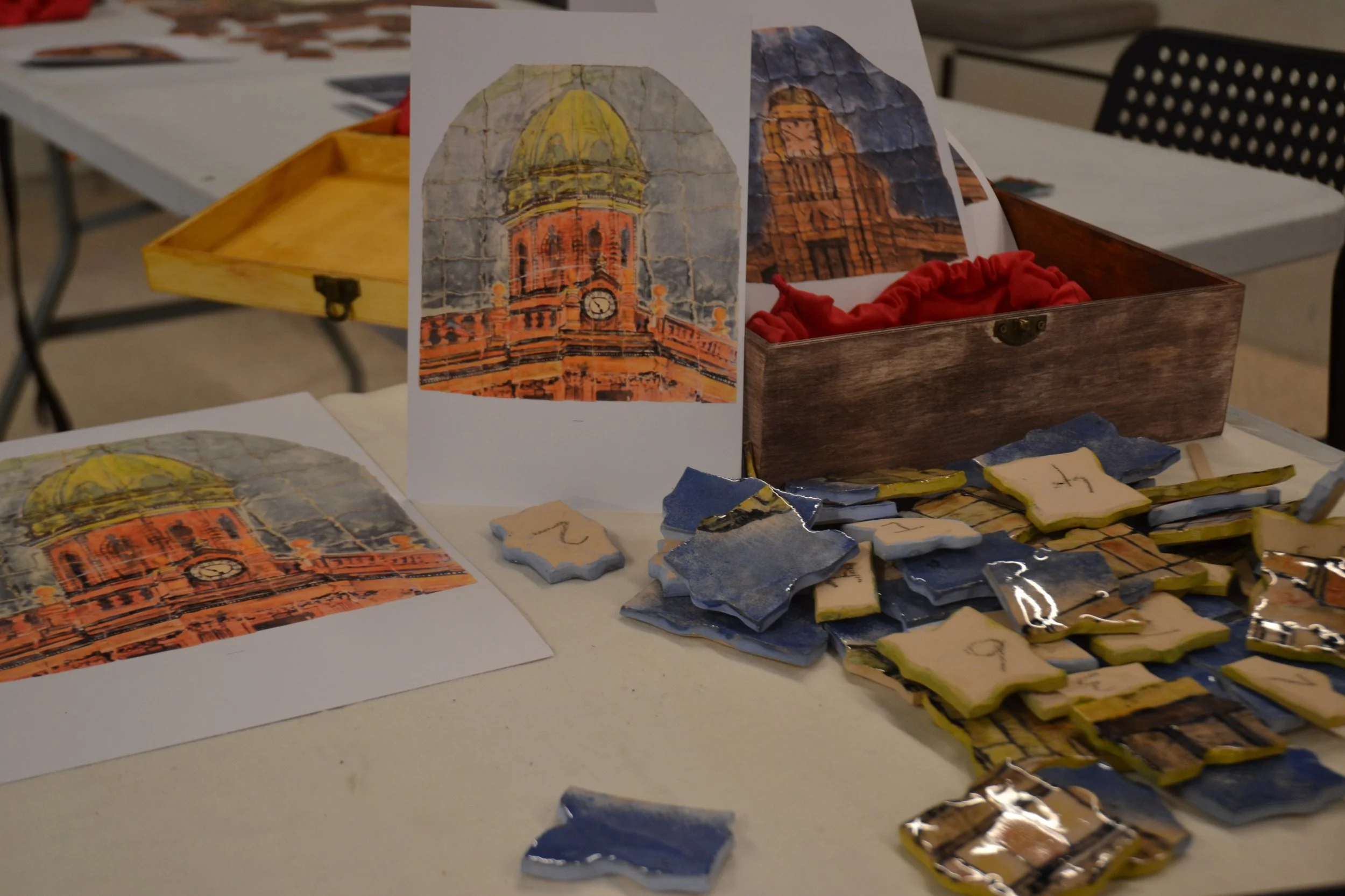 A table at an art market displaying printed images of a historic clock tower and scattered handmade ceramic tiles, some with handwritten numbers.