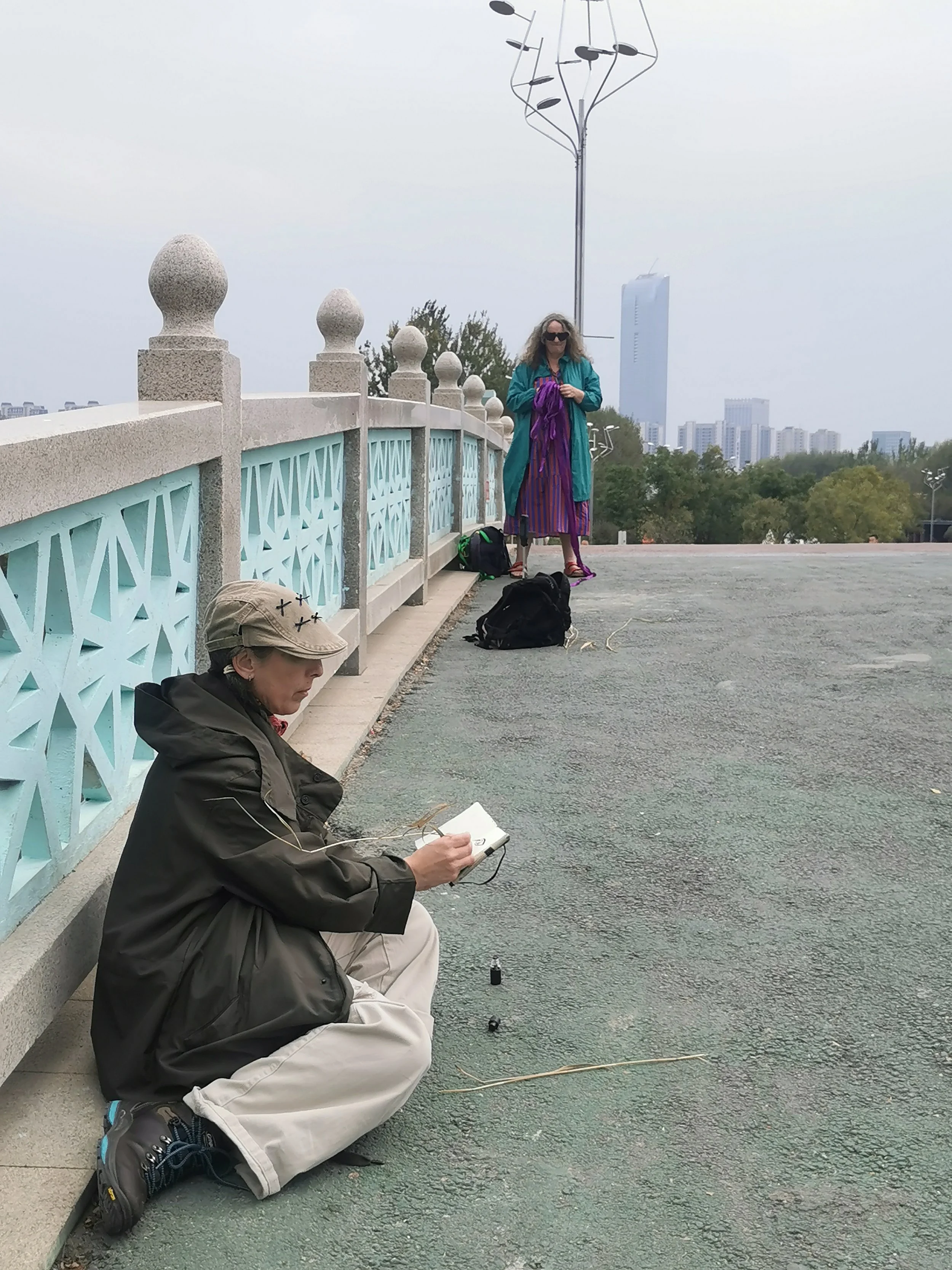 A woman sitting on the ground drawing in a book, two backpacks nearby, and a person standing in the background with a city skyline and trees behind her.
