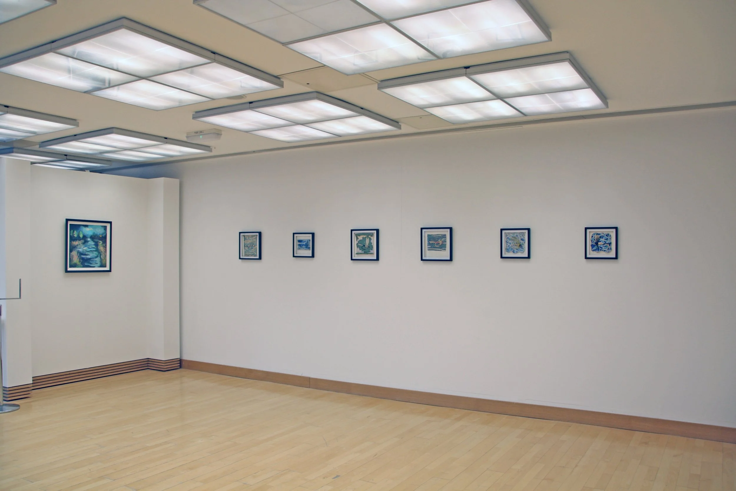 An art gallery with framed paintings on a white wall, a wooden floor, and ceiling lights.