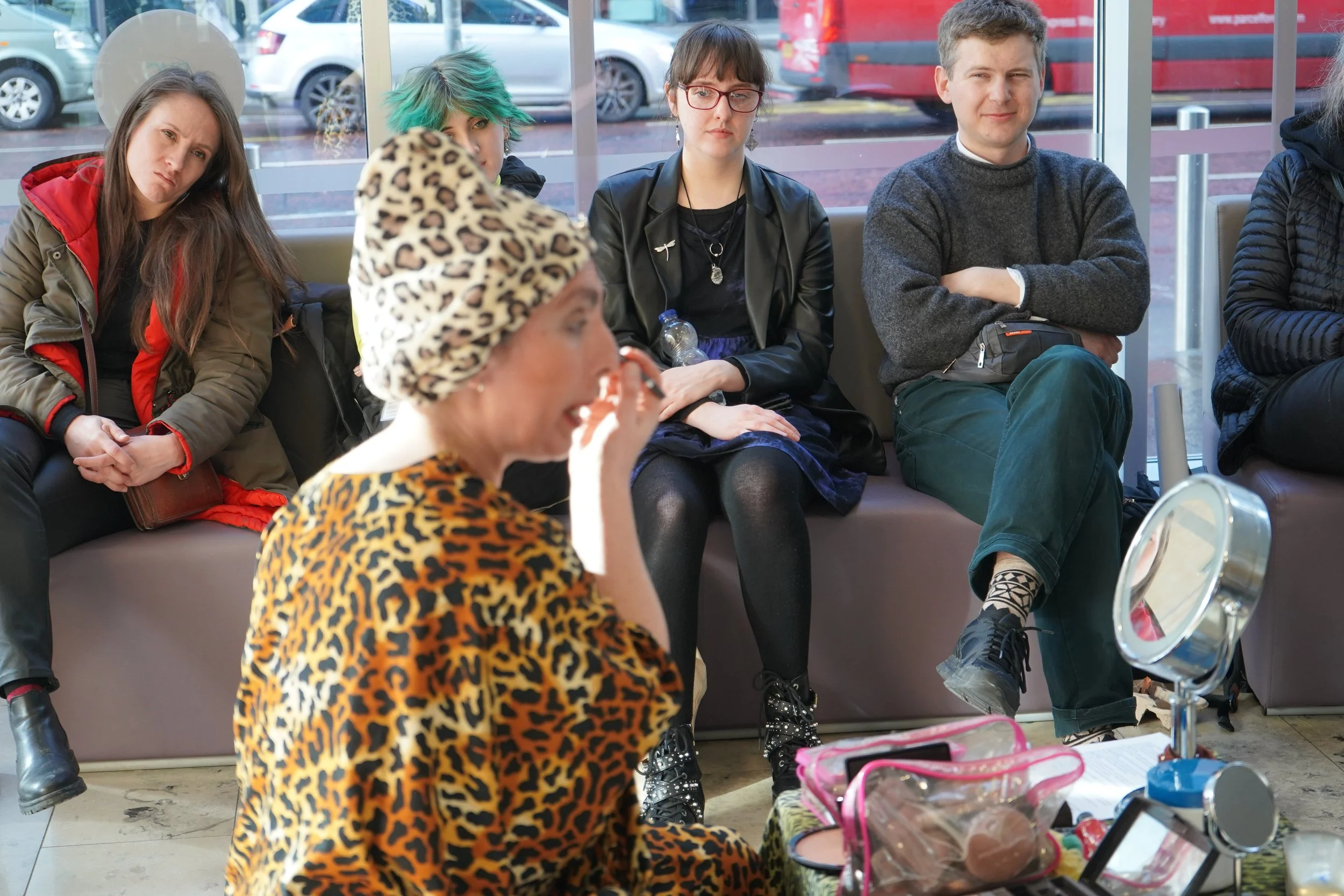 Group of five young adults sitting in a waiting area, some looking at the camera, with cars visible outside through large windows behind them.