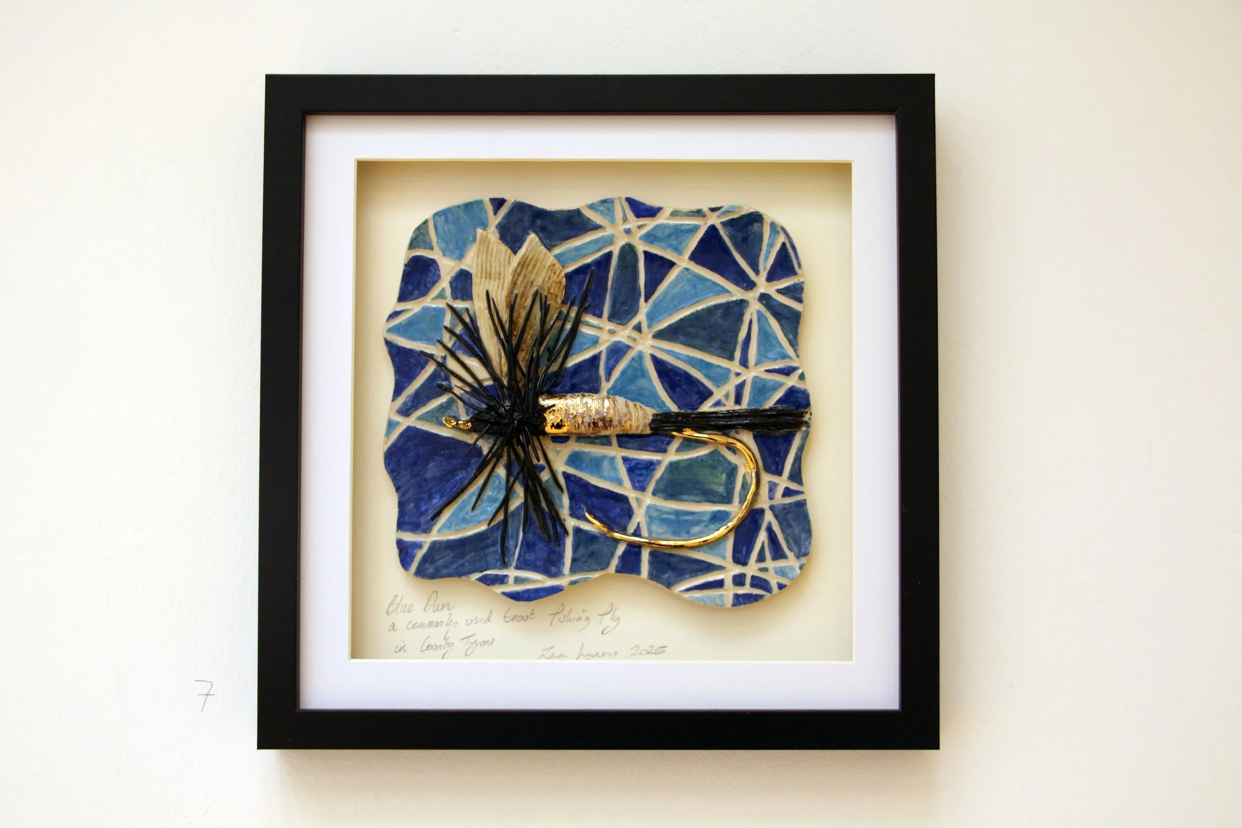 A framed mixed media art piece depicting a blue butterfly with black antennae and a gold-colored body, set against a mosaic background of blue and cream geometric tiles.