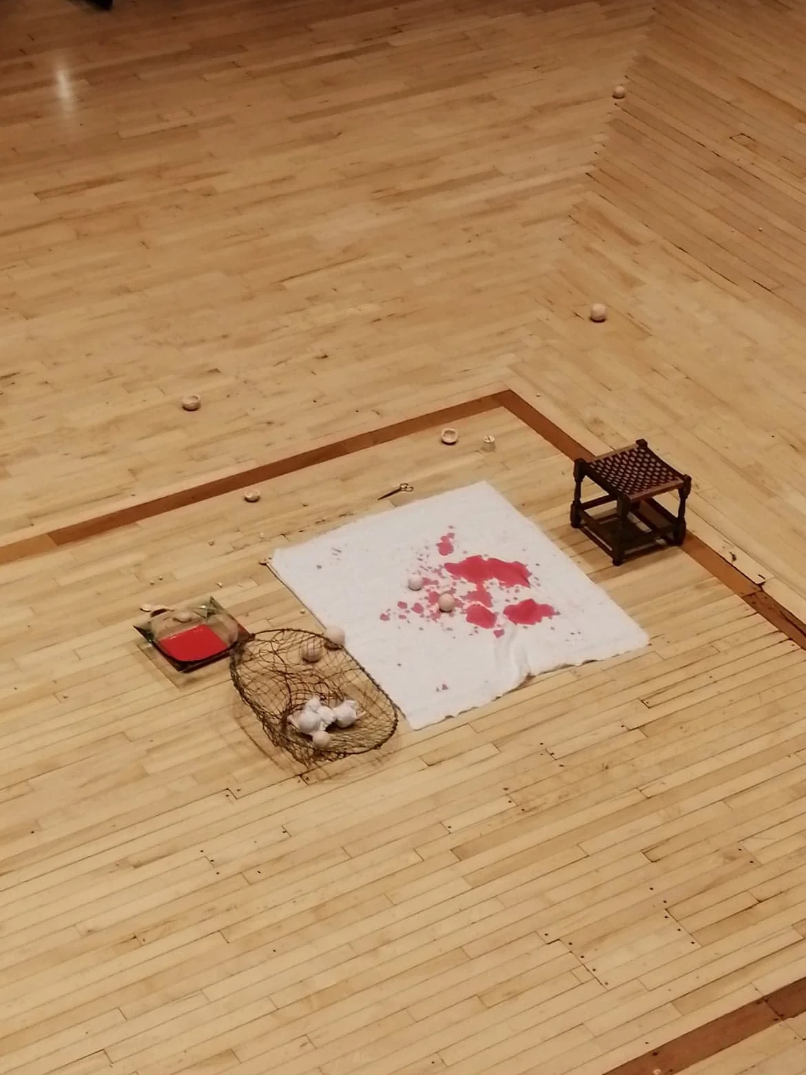 A badminton court with a white towel covered in red paint, scattered around balls, a small wooden stool, a net with balls inside, and a square container with red paint.