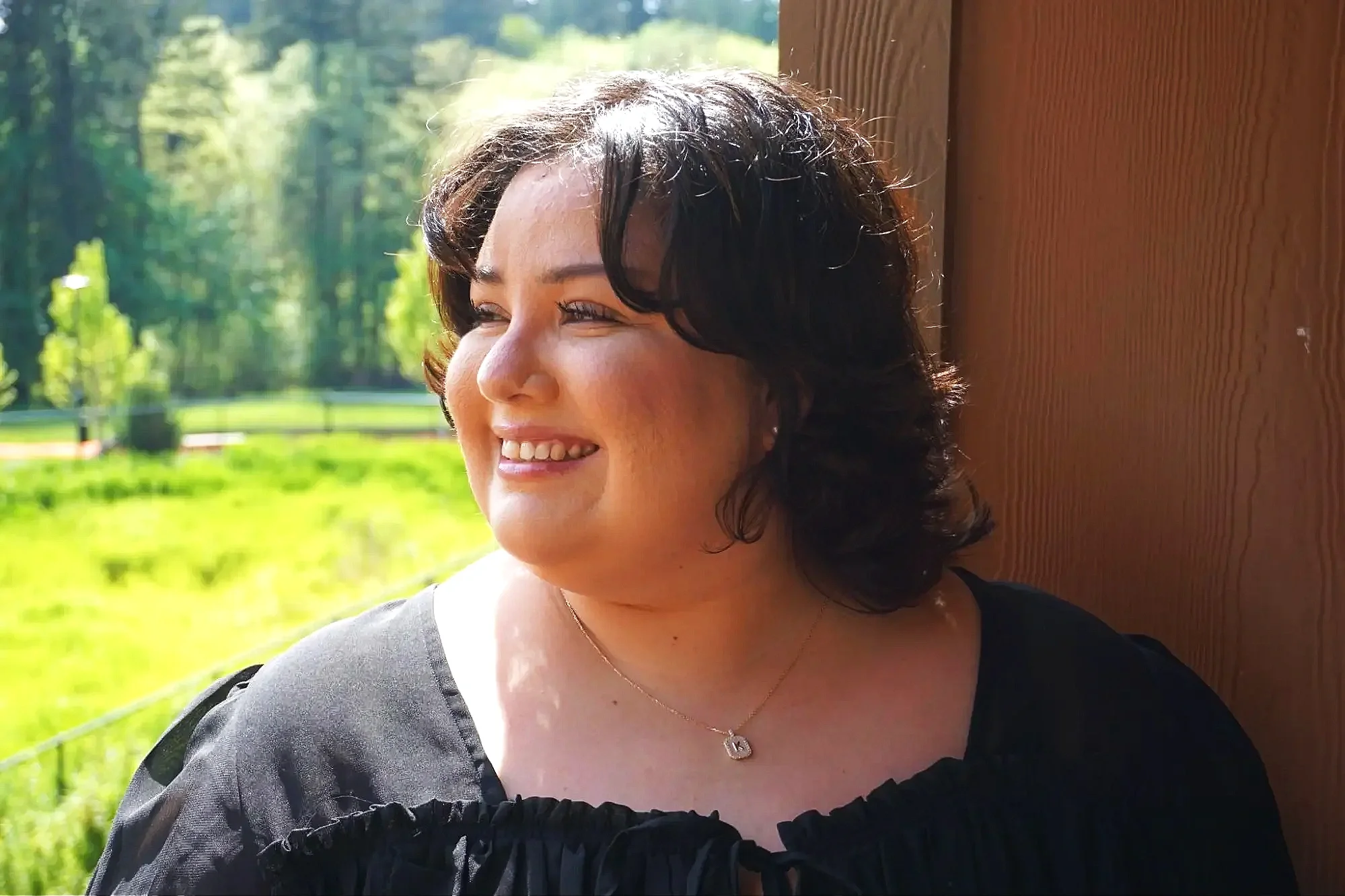 Karla smiling with short dark hair wearing a black top and a gold necklace with a heart pendant, looking to the left, outdoors with green trees and grass in the background.