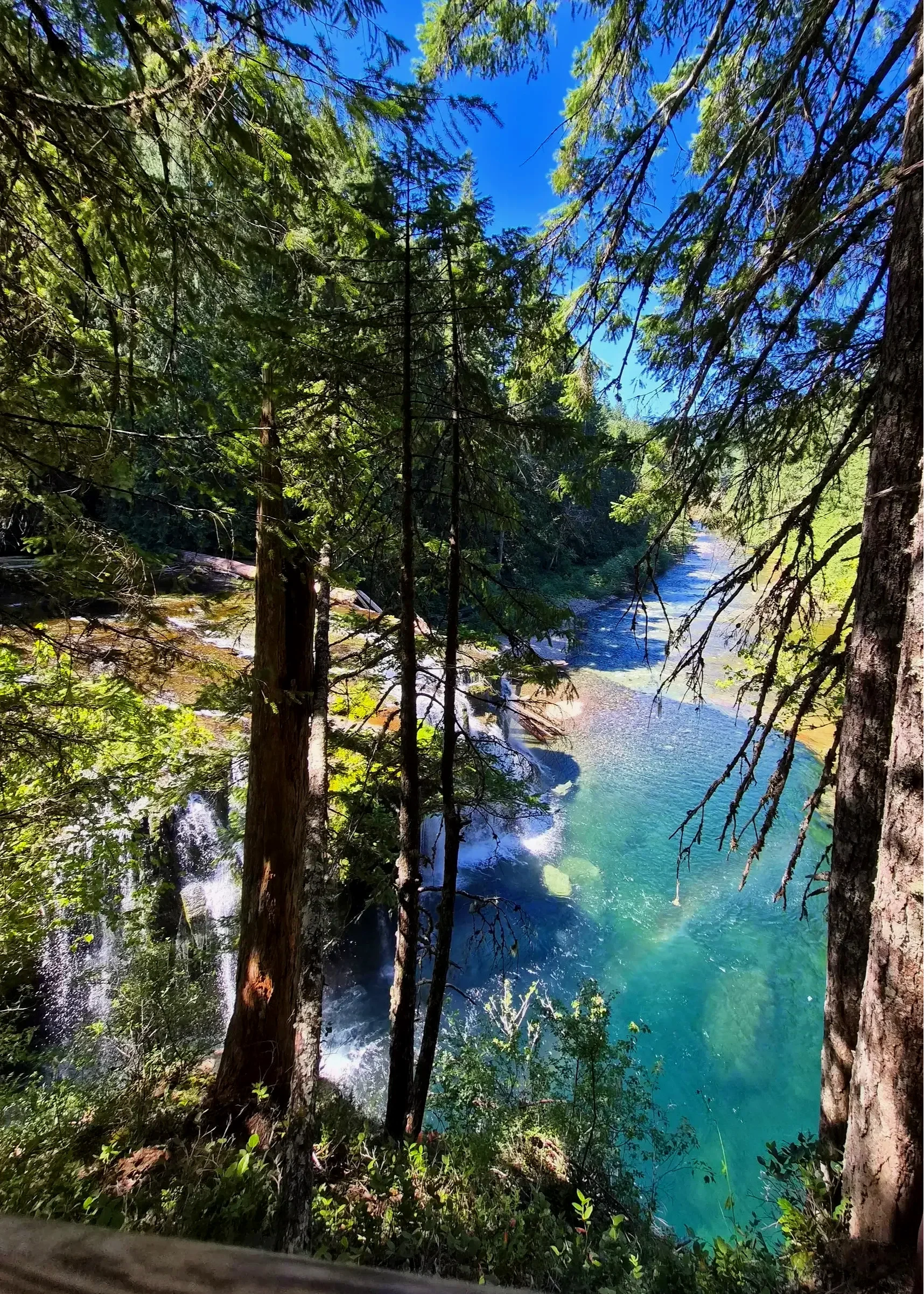 Vista desde la cima de una cascada, con árboles a ambos lados y un pequeño arcoíris sobre el agua azul y cristalina.