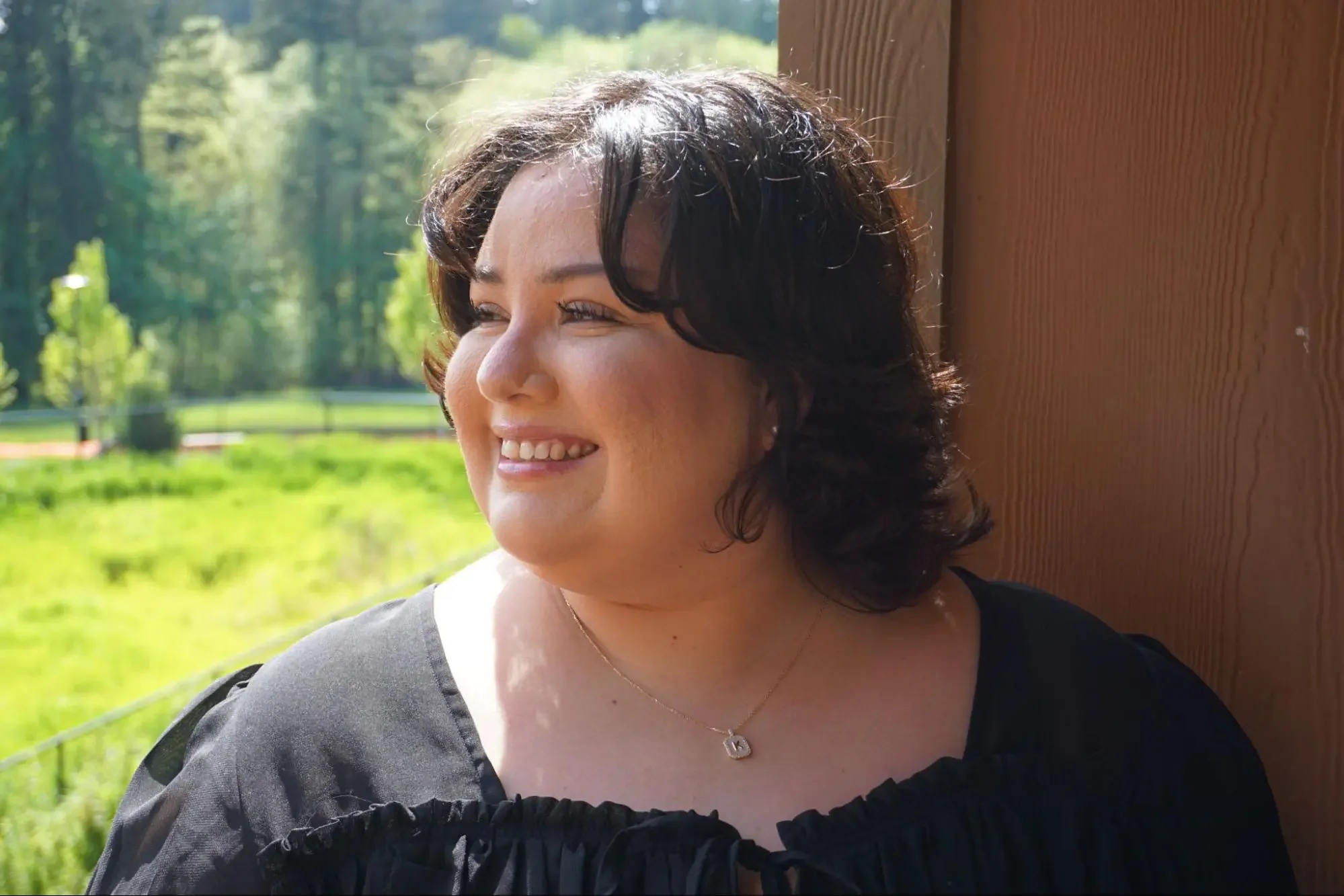 Karla sonriendo, con el cabello corto y oscuro, vistiendo una blusa negra y un collar dorado con un colgante en forma de corazón; mira hacia la izquierda y se encuentra al aire libre, con árboles verdes y césped de fondo.