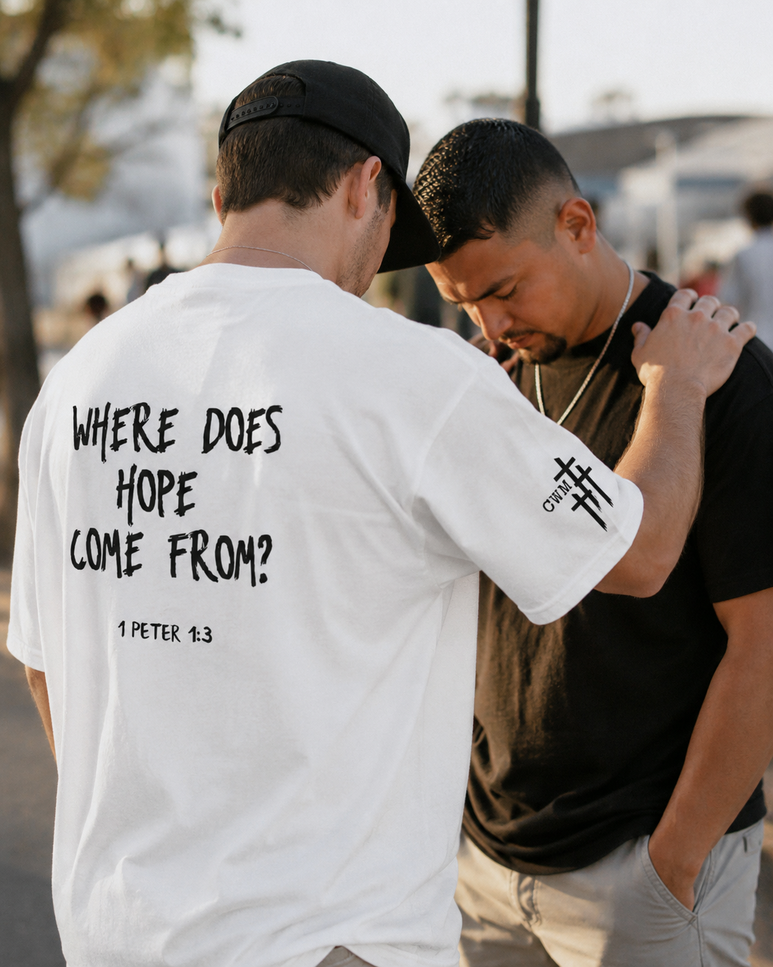 Two men are praying together outdoors, one wearing a white shirt with the Bible verse 1 Peter 1:3 written on the back, and the other wearing a black shirt, with their heads bowed and one touching the other’s shoulder.