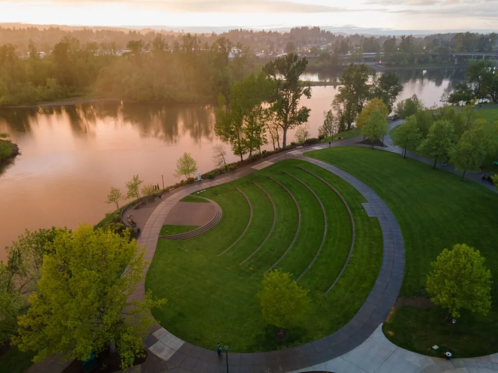 A scenic park with a curved amphitheater and paved walkways beside a river, surrounded by trees with some in autumn foliage, during sunset.