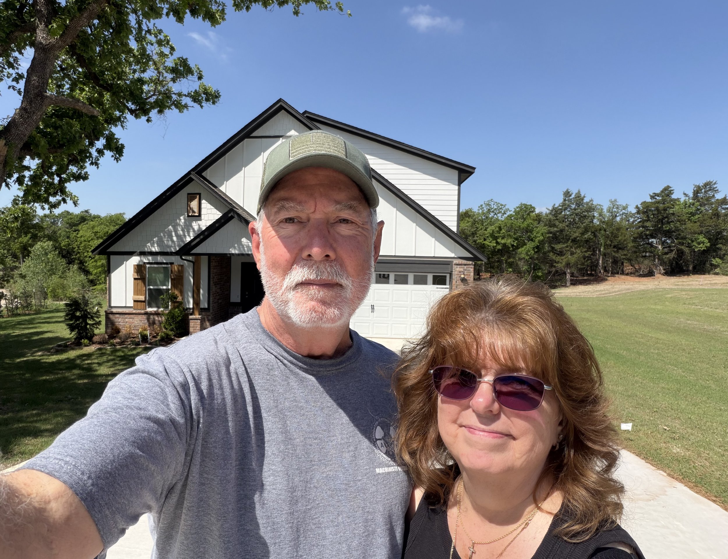 Close-up selfie of an older man and woman outdoors in front of a house, with trees and a blue sky in the background.