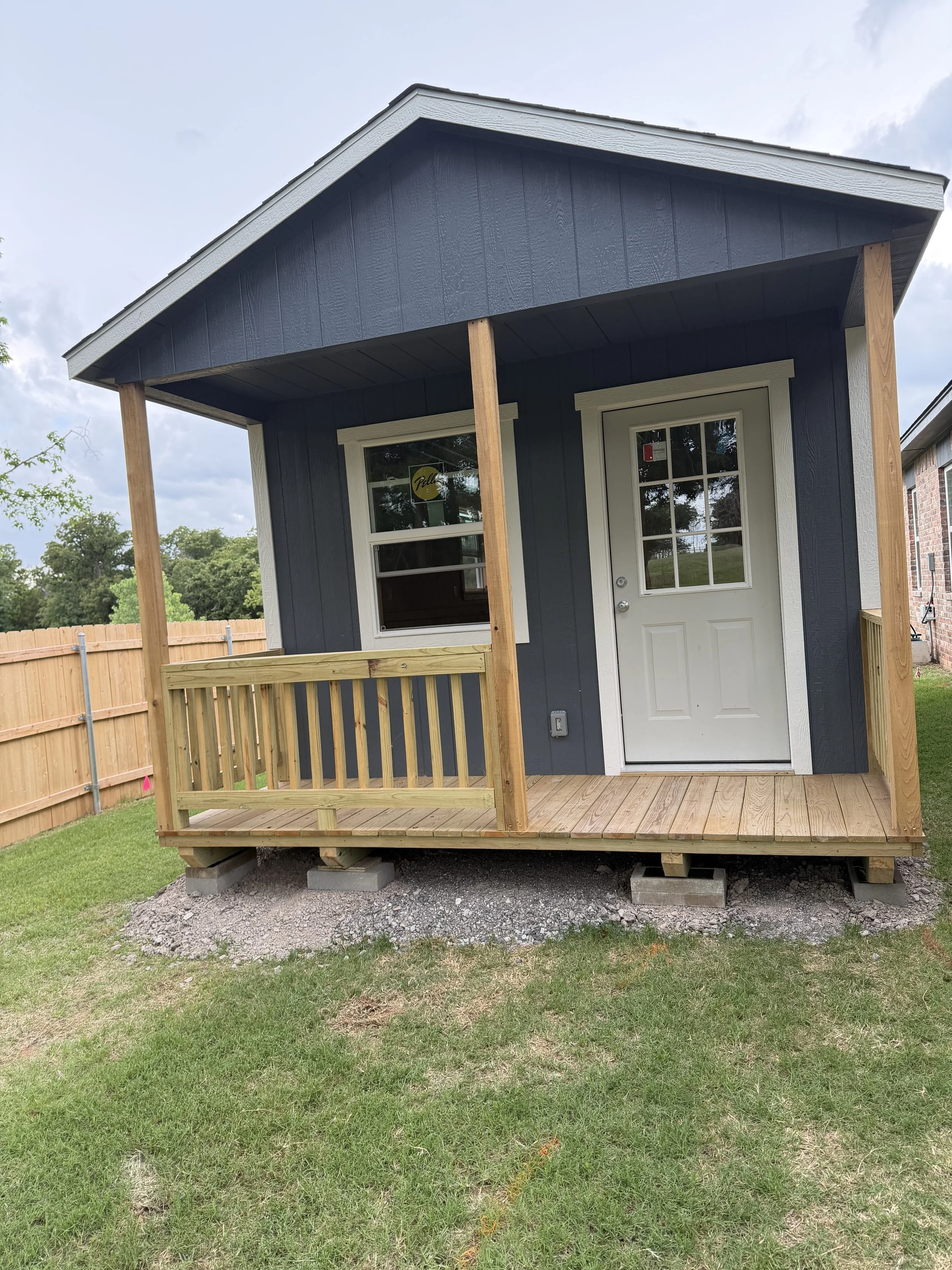 A small wooden porch with a white door, a window, and a dark blue exterior, supported by four wooden posts, with a grassy lawn and a wooden fence in the background.