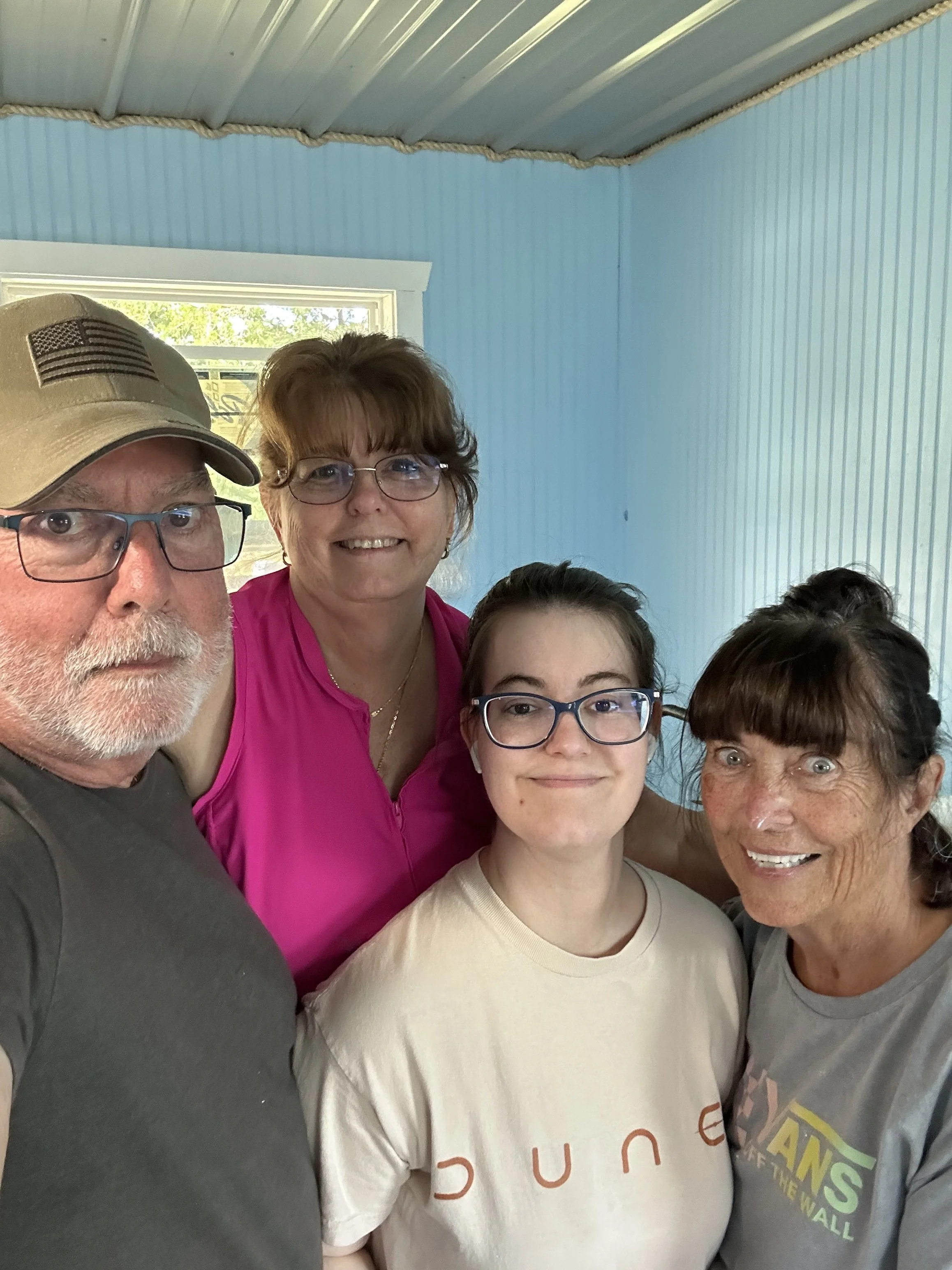 A group of five people taking a selfie inside a room with light blue paneled walls and a window showing green foliage outside. The group includes two older women, a man wearing a beige cap, and two younger women with glasses.
