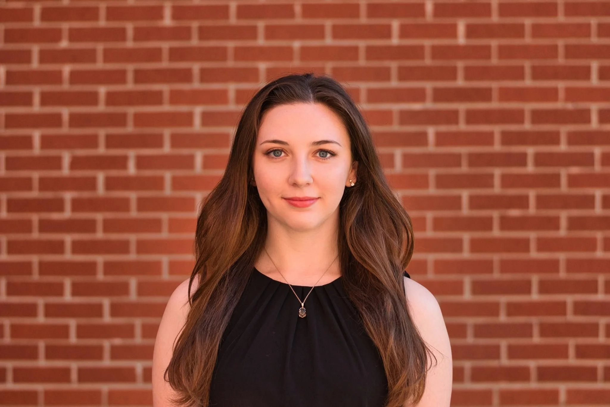 A young woman with long brown hair and blue eyes posing in front of a brick wall. She is wearing a black sleeveless top, earrings, and a necklace.