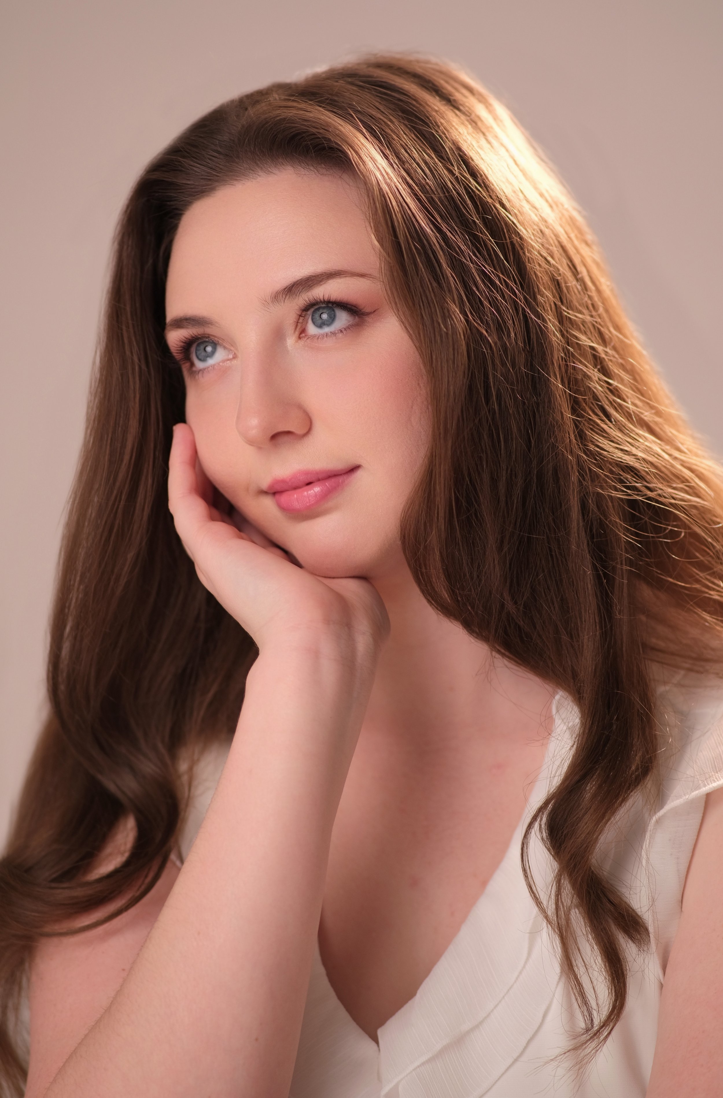 A young woman with long, wavy brown hair and blue eyes resting her face on her hand, looking contemplative.