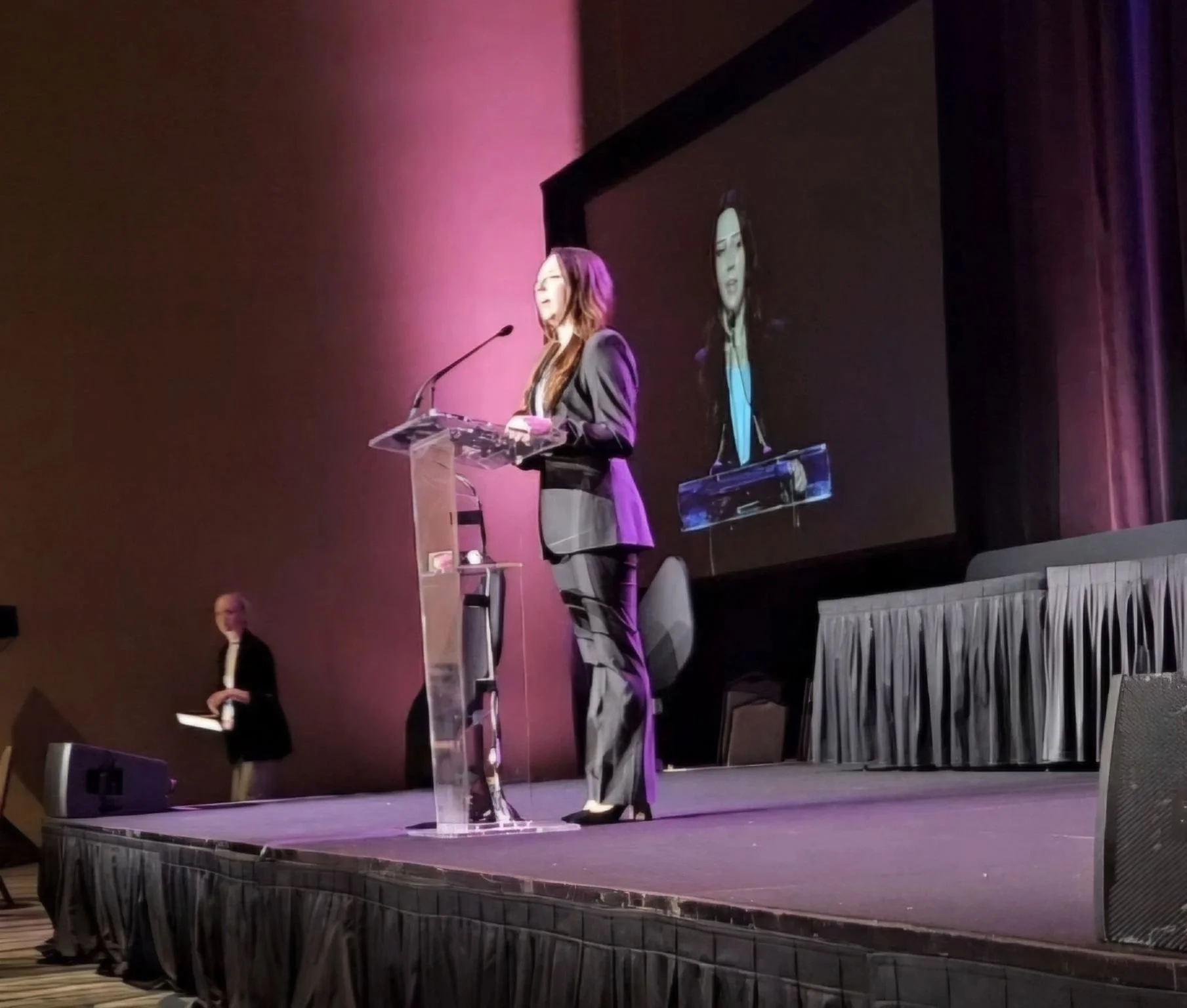 A woman in a dark blazer and pants stands at a clear podium on stage, speaking into a microphone. Her reflection is visible on a large screen behind her. Another person in a black suit with white shirt stands in the background, holding an object, perhaps a folder or tablet. The stage has purple and pink lighting, with a skirted platform and some equipment in the foreground.